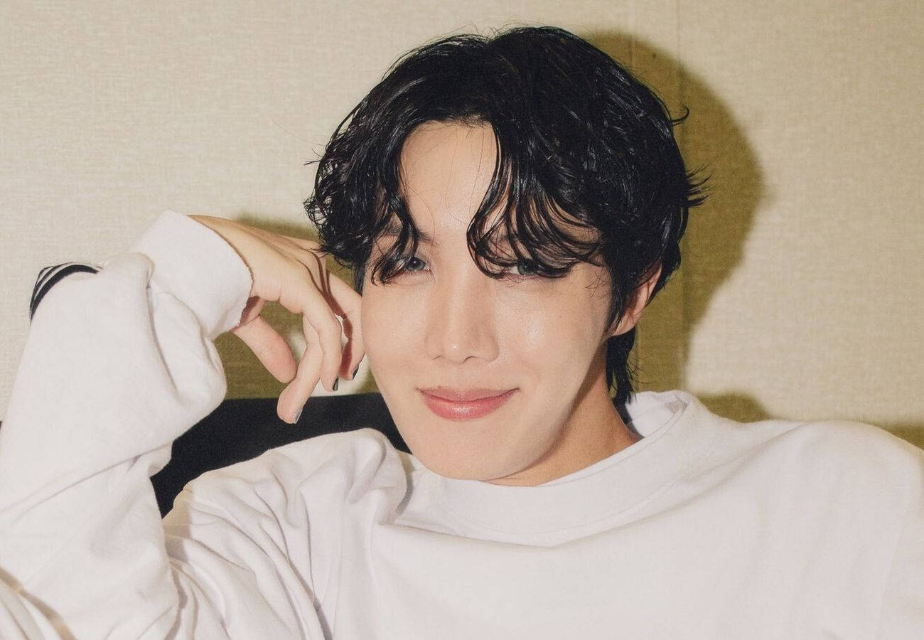 可愛微笑的 J-Hope 壁紙 — 一位深色波浪髮的年輕男子，身穿白色衛衣，將手放在臉旁，溫柔微笑。