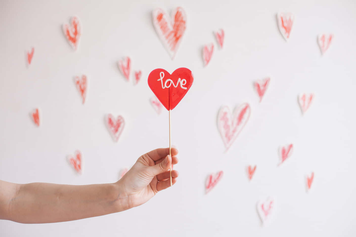 手拿著一個紅色心形，上面寫著 love，附在棍子上，對著一面裝飾著心形的牆壁。