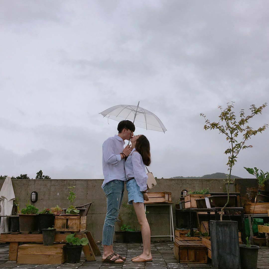 可愛情侶撐傘於天台咖啡廳 壁紙 — 一對情侶在雨中的屋頂花園裡，在透明雨傘下接吻，周圍有盆栽植物和木箱。