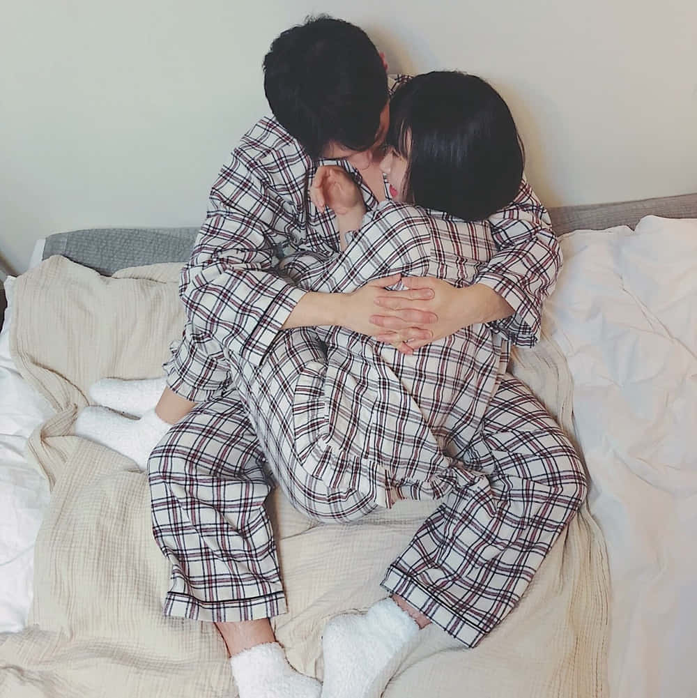 可愛時尚情侶床上擁抱照 壁紙 — 一對穿著相配格仔睡衣的情侶在鋪有柔軟米色床單的舒適床上溫柔擁抱。