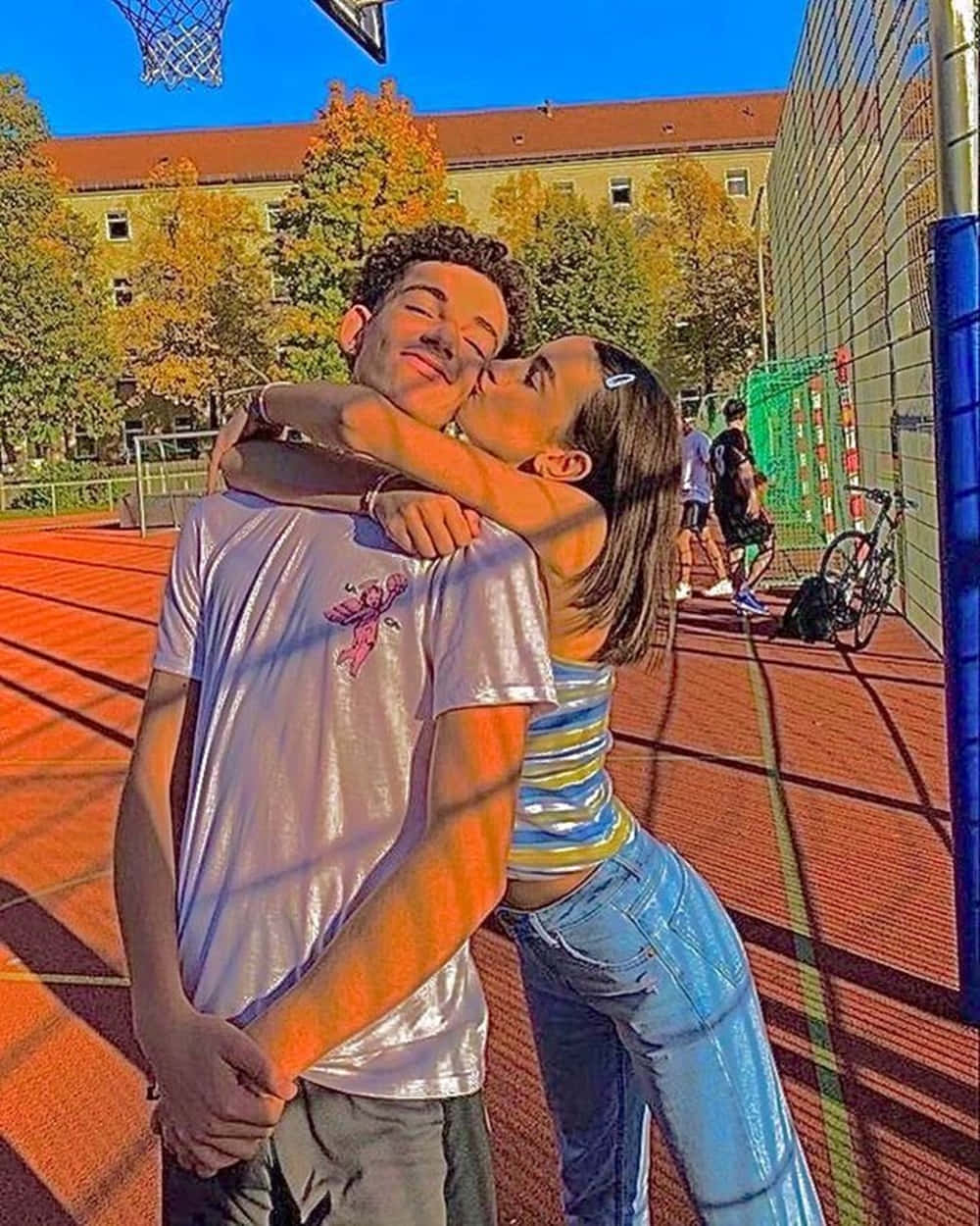 可愛時尚情侶親吻頰部照片 壁紙 — 一對年輕情侶在陽光明媚的戶外籃球場上深情接吻，周圍是秋天的樹木。