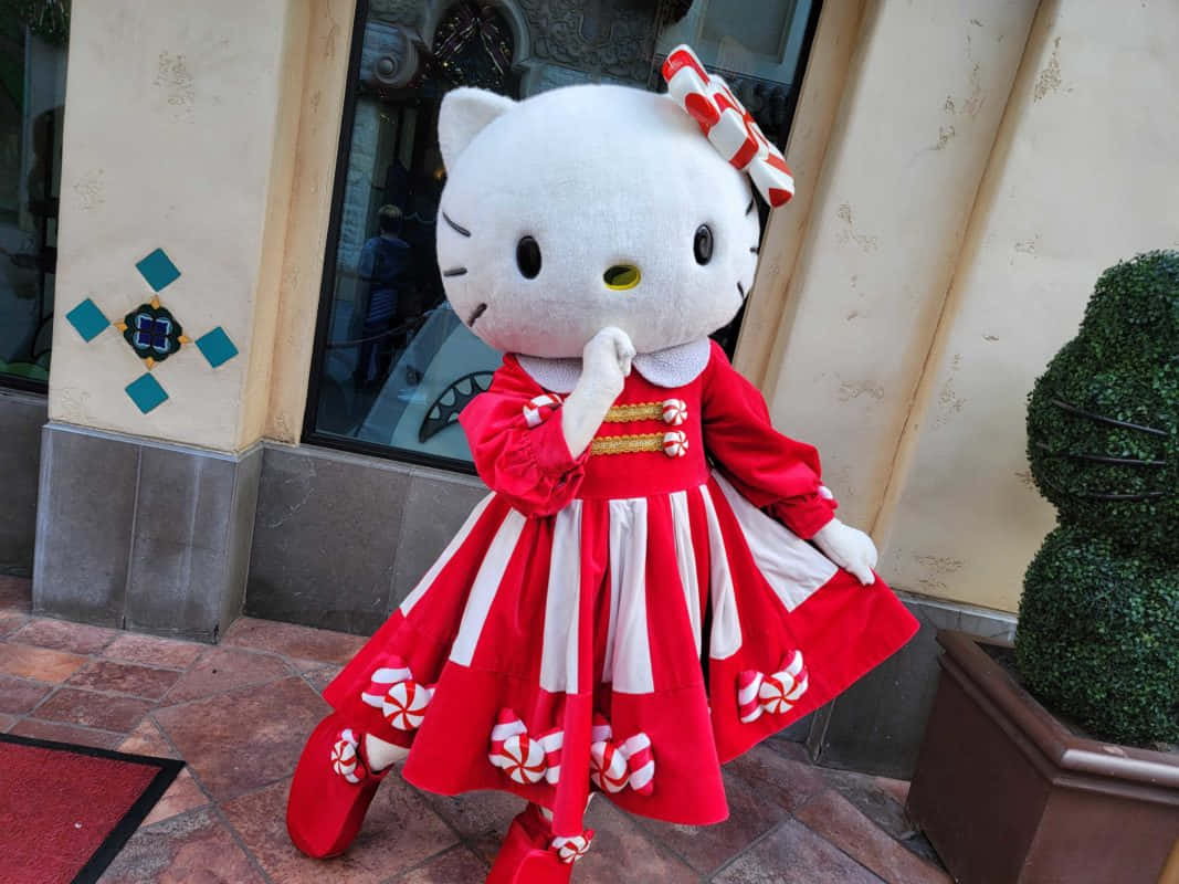 可愛有趣的 Hello Kitty 藝術 壁紙 — 一個大型Hello Kitty吉祥物穿著節日紅白條紋連衣裙，站在瓷磚走道上，旁邊是一座建築物和盆栽灌木。