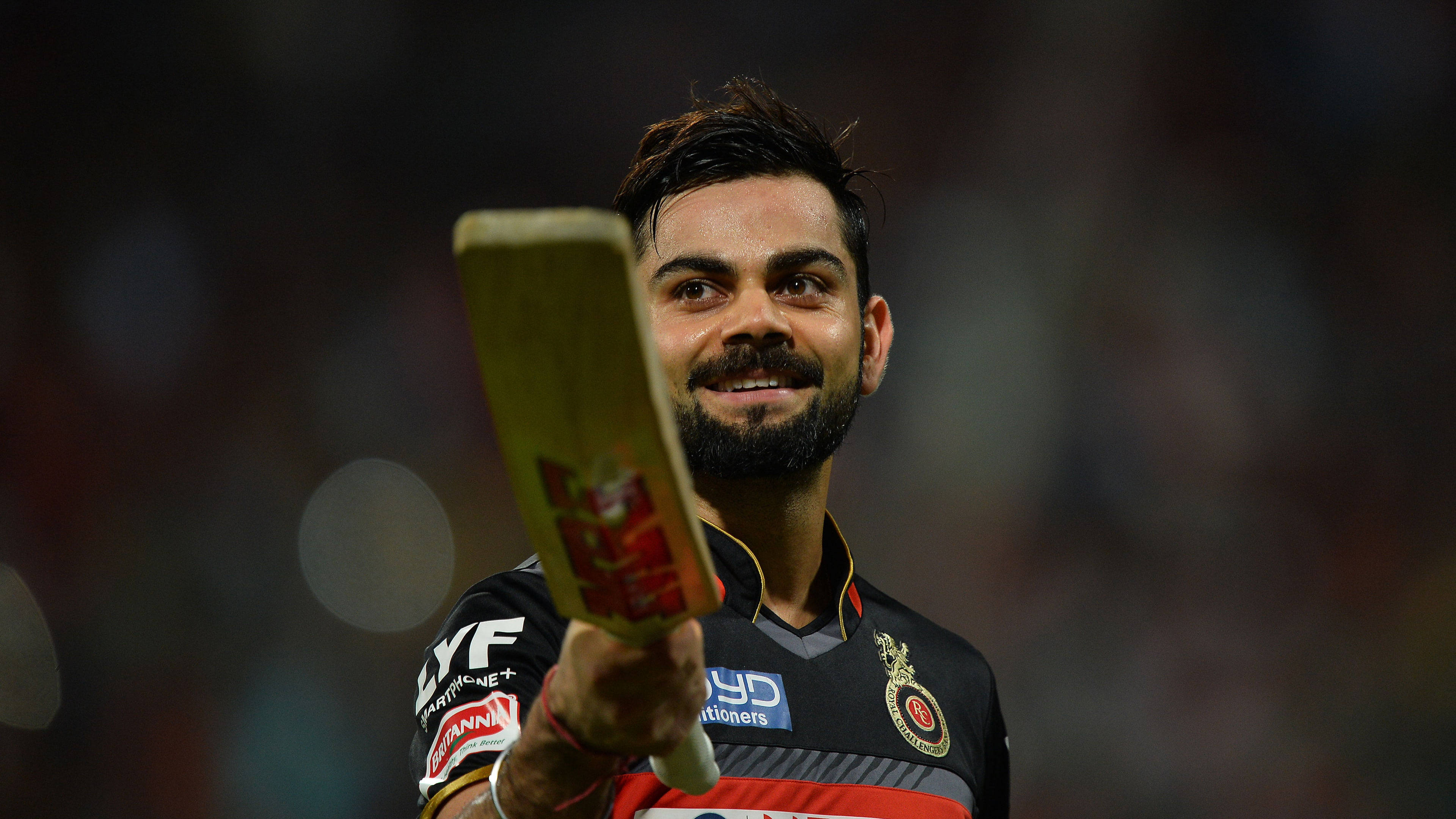 可愛板球運動員 4K 壁紙 — 板球運動員 Virat Kohli 在夜間比賽中自豪地微笑，手持球棒，在體育場燈光下。