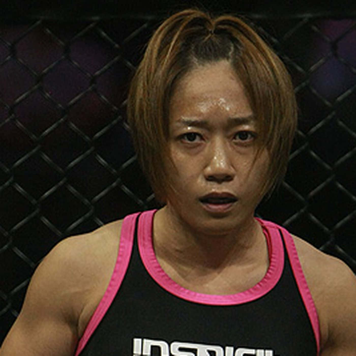 可愛梅古美·藤井近距離特寫 壁紙 — 一位專注的女戰士穿著黑色粉紅邊背心，在昏暗的MMA八角籠內凝視著。