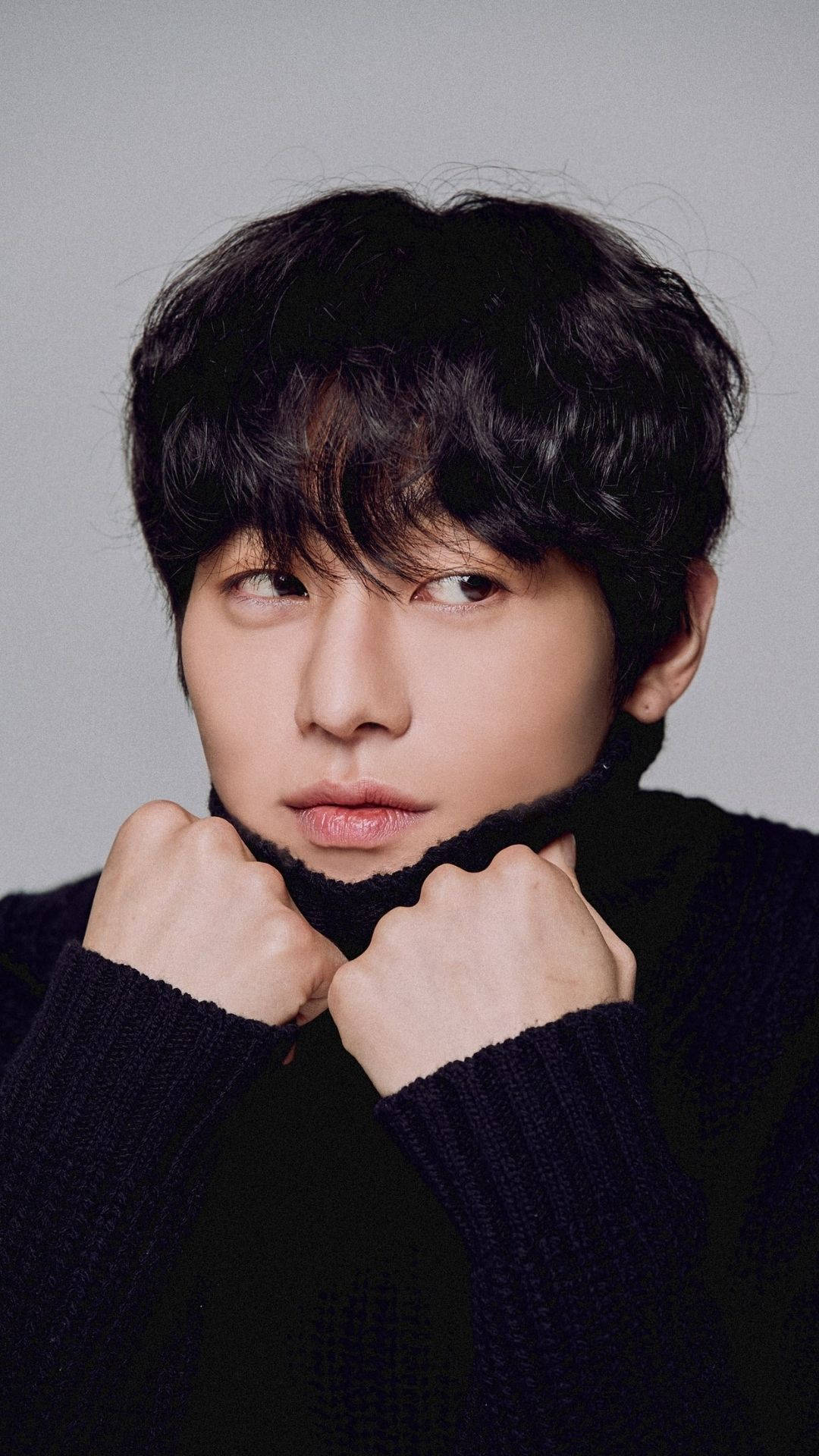 可愛演員安孝燮 壁紙 — 一位年輕男子留著深色波浪頭髮，在純灰色背景前抓住黑色針織毛衣的領口。