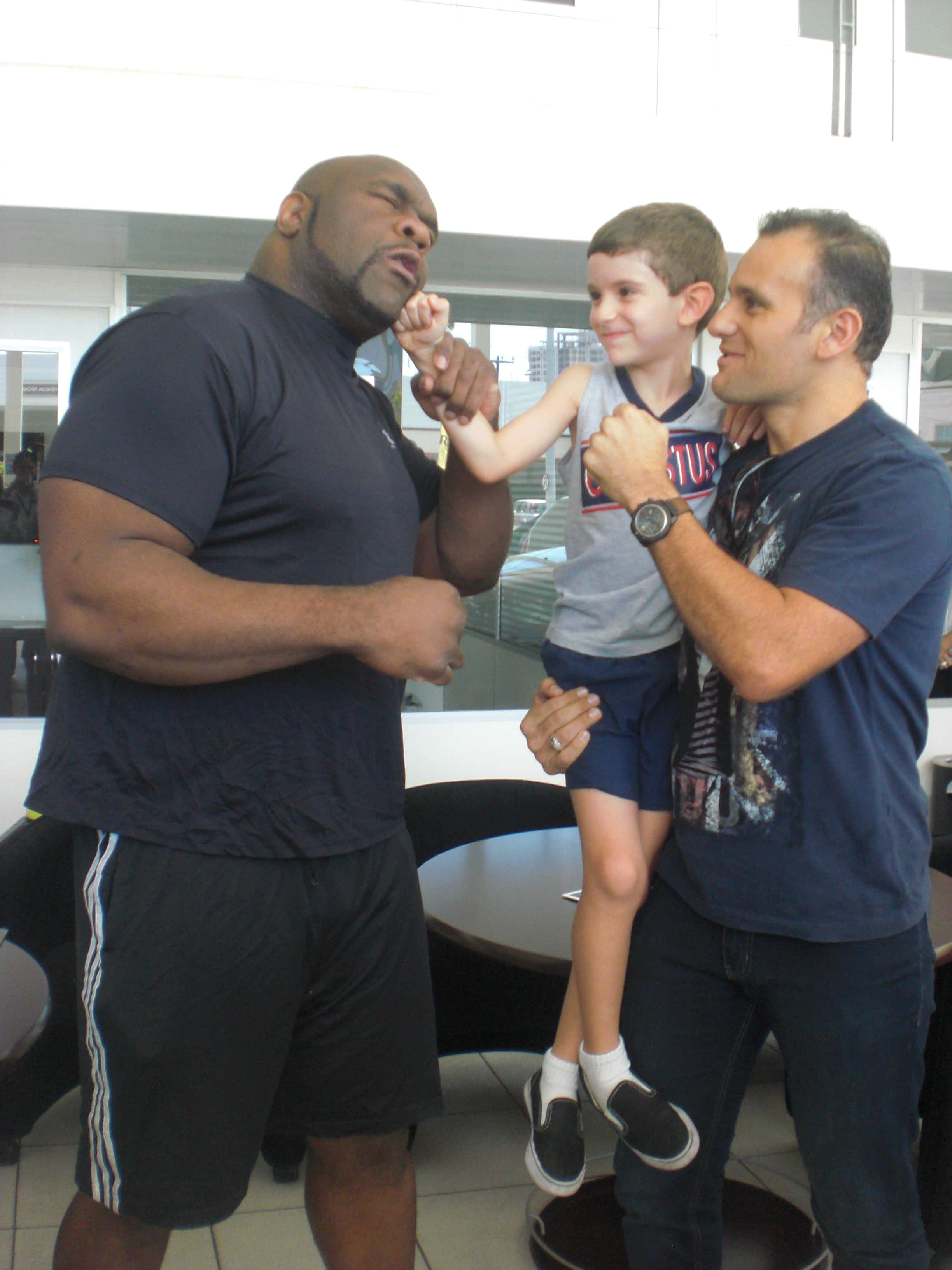 可愛的 Bob Sapp 與他的粉絲 壁紙 — 一個肌肉發達的男人與另一個男人抱著的小孩在現代室內環境中嬉戲互動。