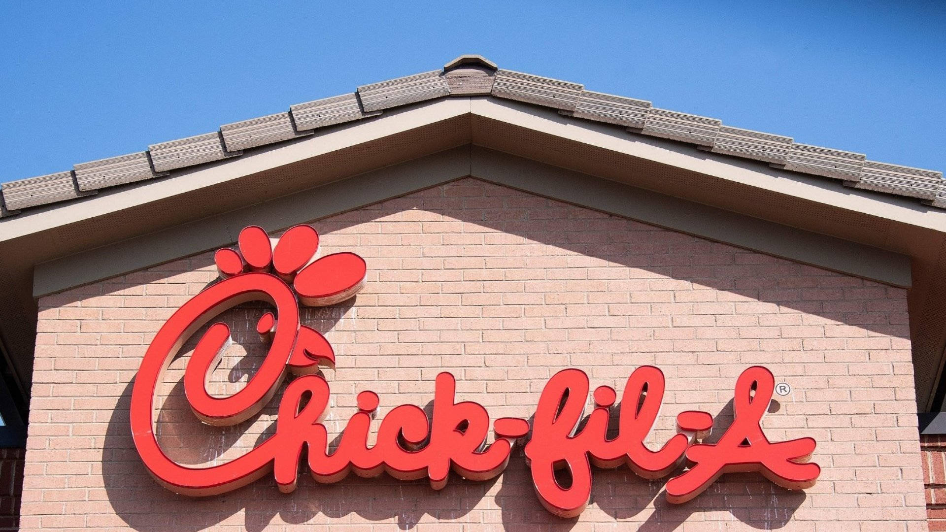 可愛的 Chick Fil A 標誌招牌 壁紙 — 大膽的紅色Chick-fil-A標誌安裝在磚砌建築上，背景是晴朗的藍天。