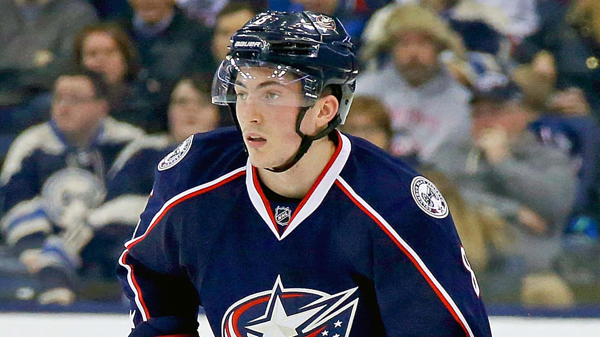 可愛的 Zachary Werenski 4K 壁紙 — 一名身穿哥倫布藍夾克球衣的專注冰球運動員在冰上凝視前方，背景觀眾模糊。