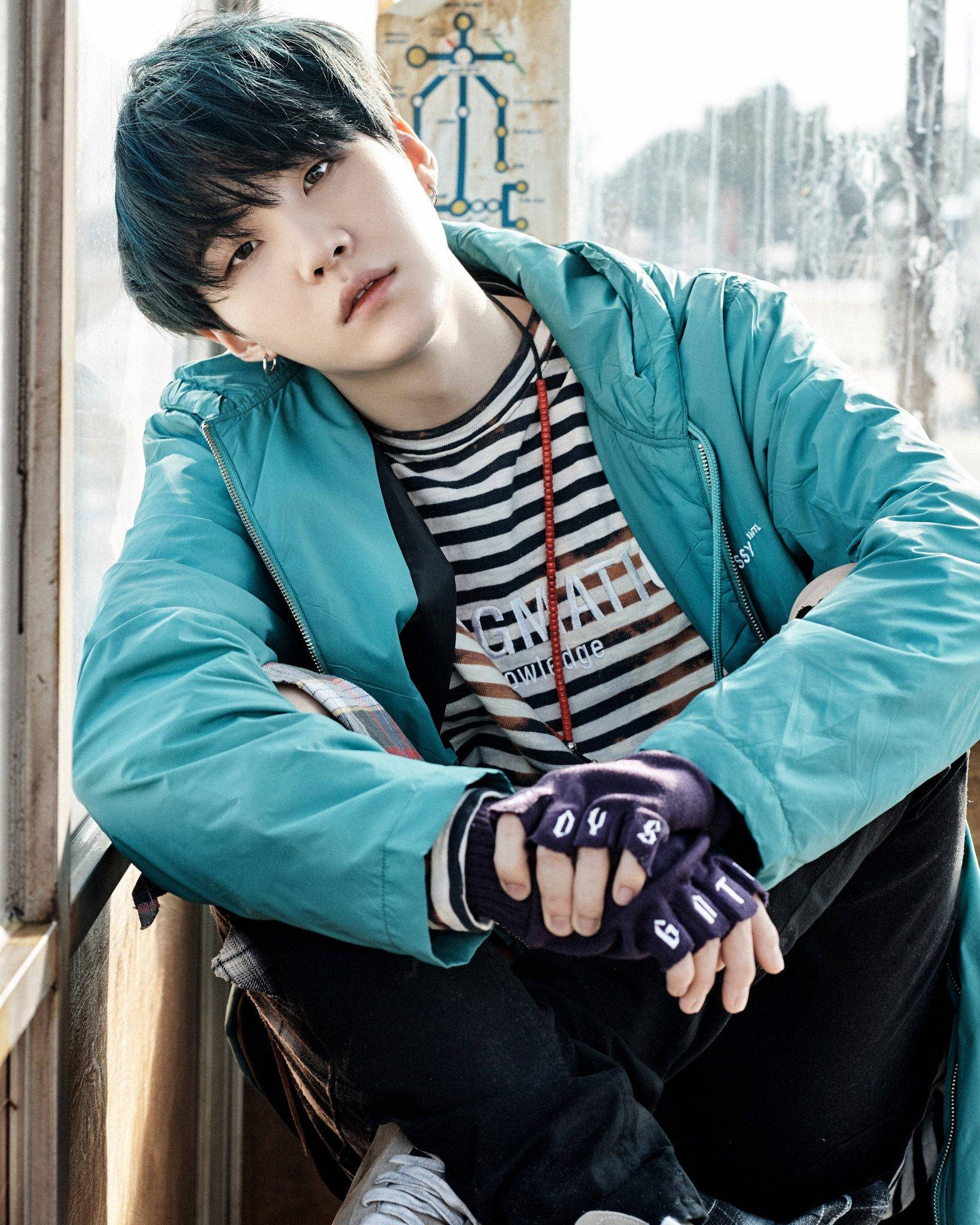 可愛的BTS SUGA穿藍色外套 壁紙 — 一名深色頭髮的年輕男子靠著窗戶，身穿青綠色外套和露指手套。