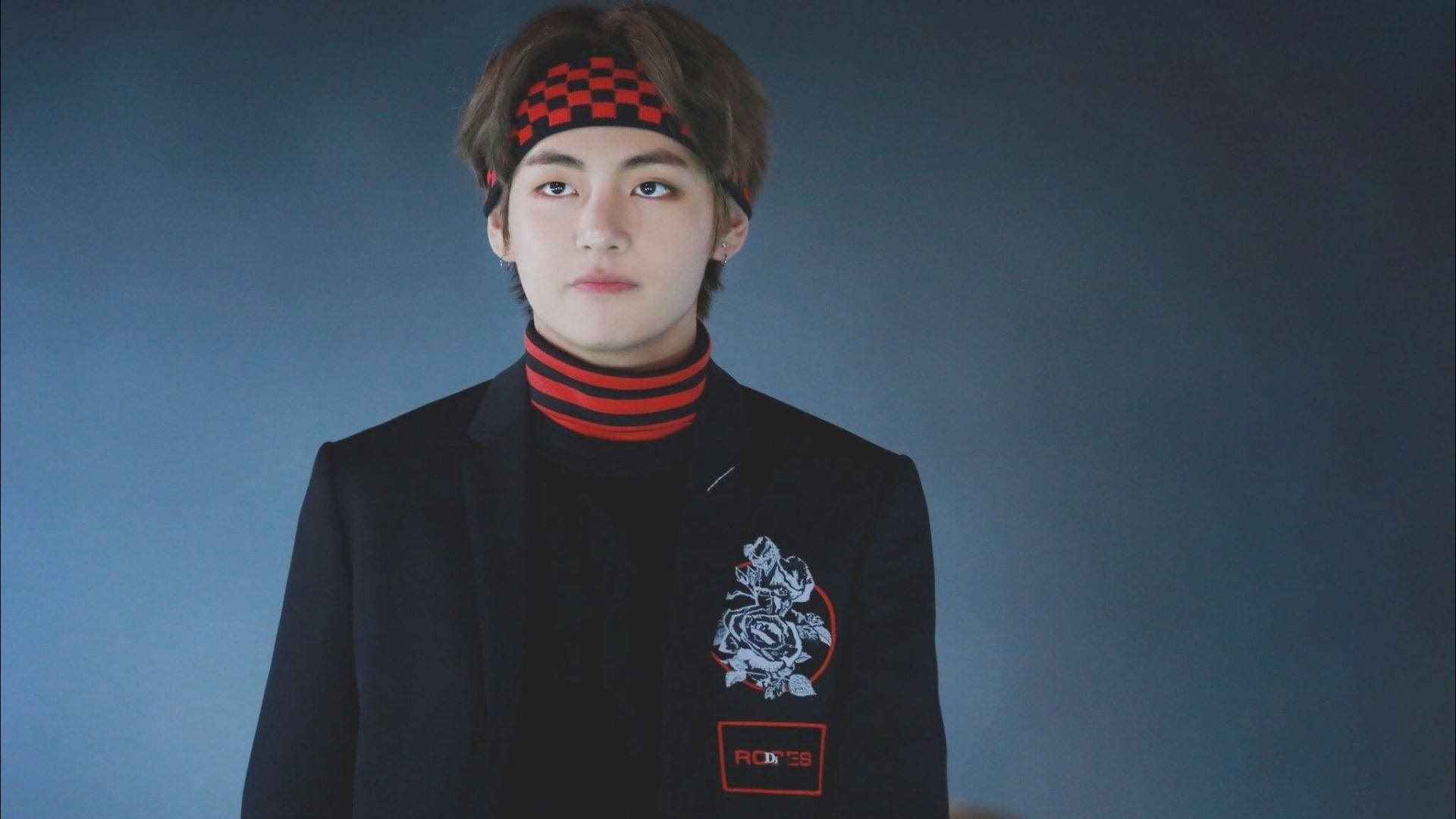 可愛的BTS V穿黑紅配色 壁紙 — 一位身穿黑色西裝外套、帶有玫瑰補丁和紅色條紋高領毛衣的年輕男子，在柔和的藍色背景下直視鏡頭。