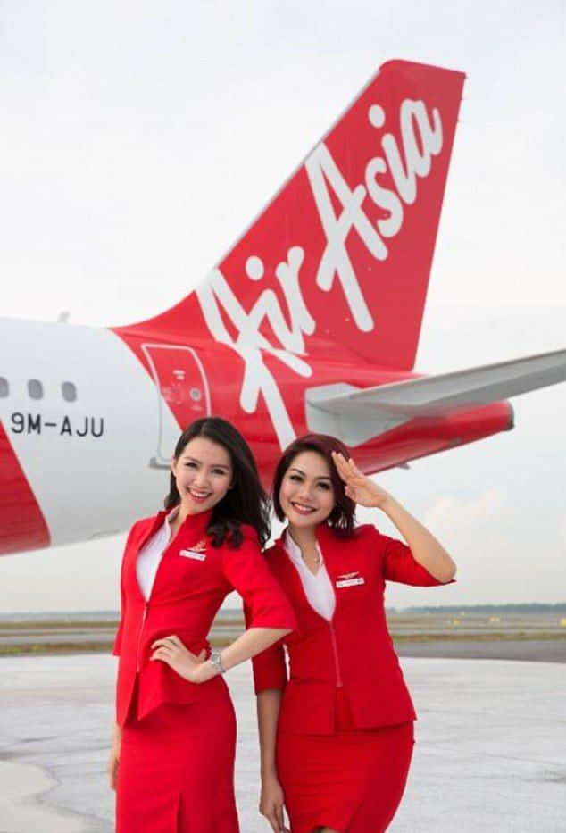 可愛的兩位空服員 壁紙 — 兩名微笑的亞洲航空空服員身穿紅色制服，在停機坪上一架紅白相間的飛機前自信地擺姿勢。