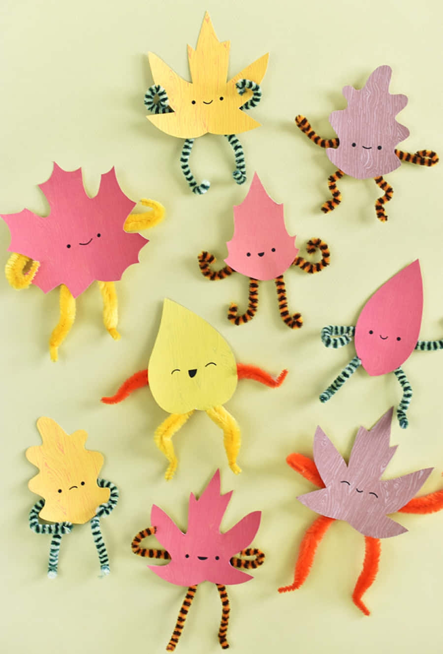 Fall Crafts 桌布 壁紙 — 可愛的紙葉角色，帶有管道清潔器四肢和微笑的臉，排列在淡黃色背景上。