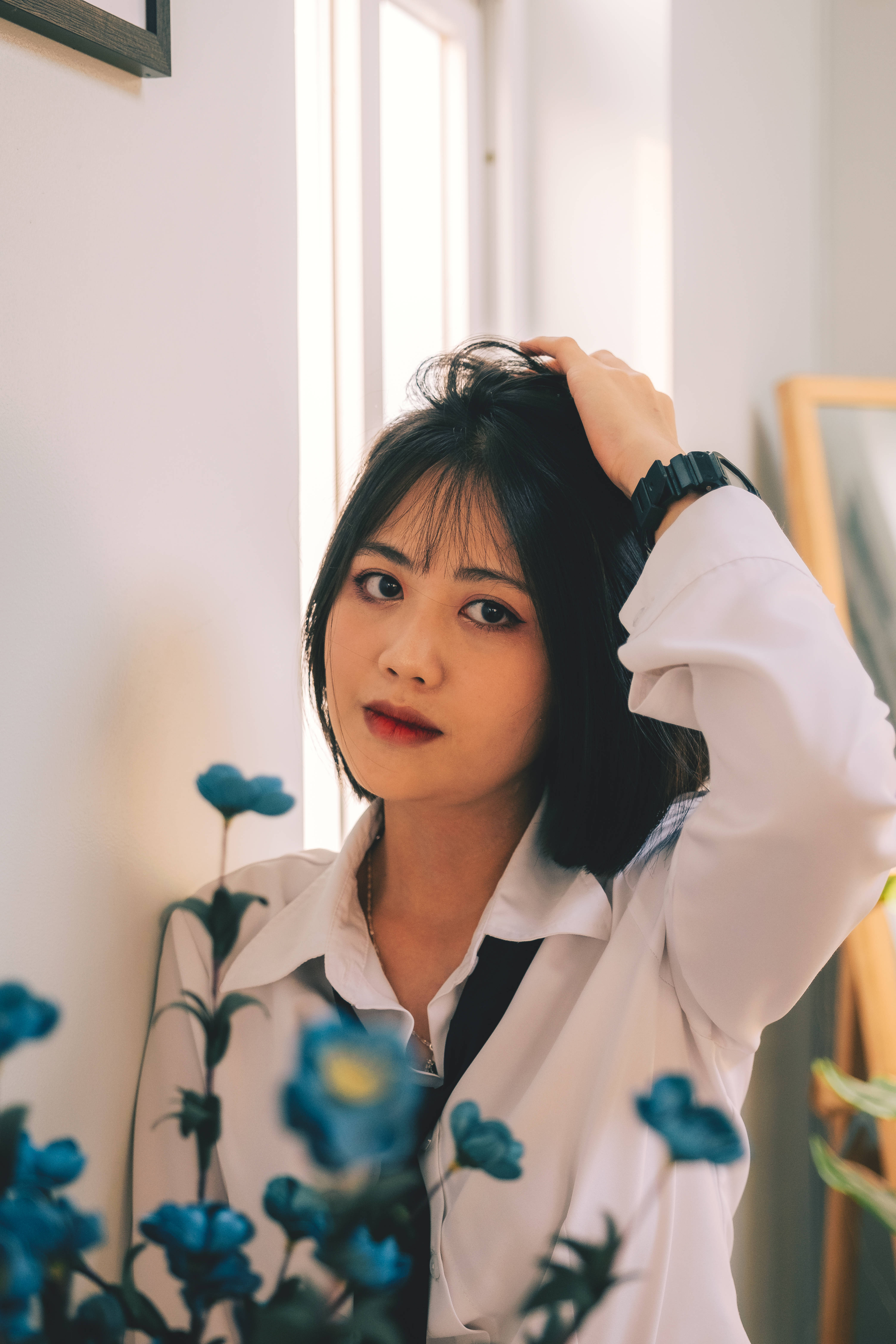 可愛的辦公室穿搭女孩 壁紙 — 一位黑髮紅唇的年輕女性，在前景有藍色花朵的窗邊輕柔地撫摸頭髮，眼神柔和。