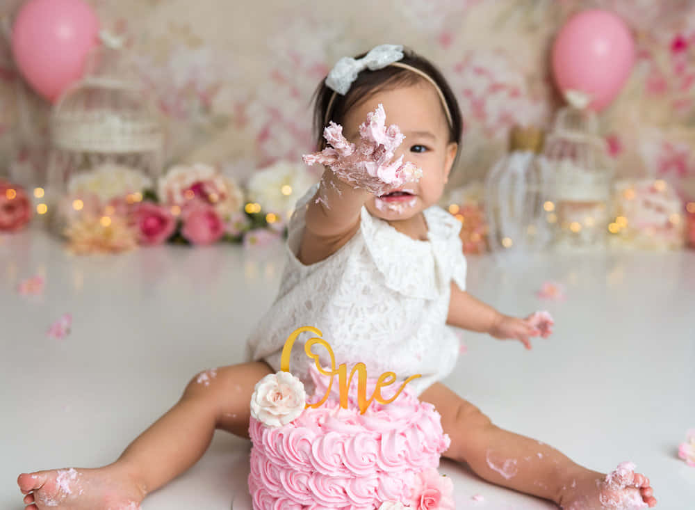 可愛的首個生日蛋糕碎屑慶祝 壁紙 — 一個快樂的女嬰穿著白色連衣裙，坐在一個覆蓋著糖霜、帶有金色「一」裝飾的粉紅色蛋糕前，她的手和臉上沾滿了糖霜。