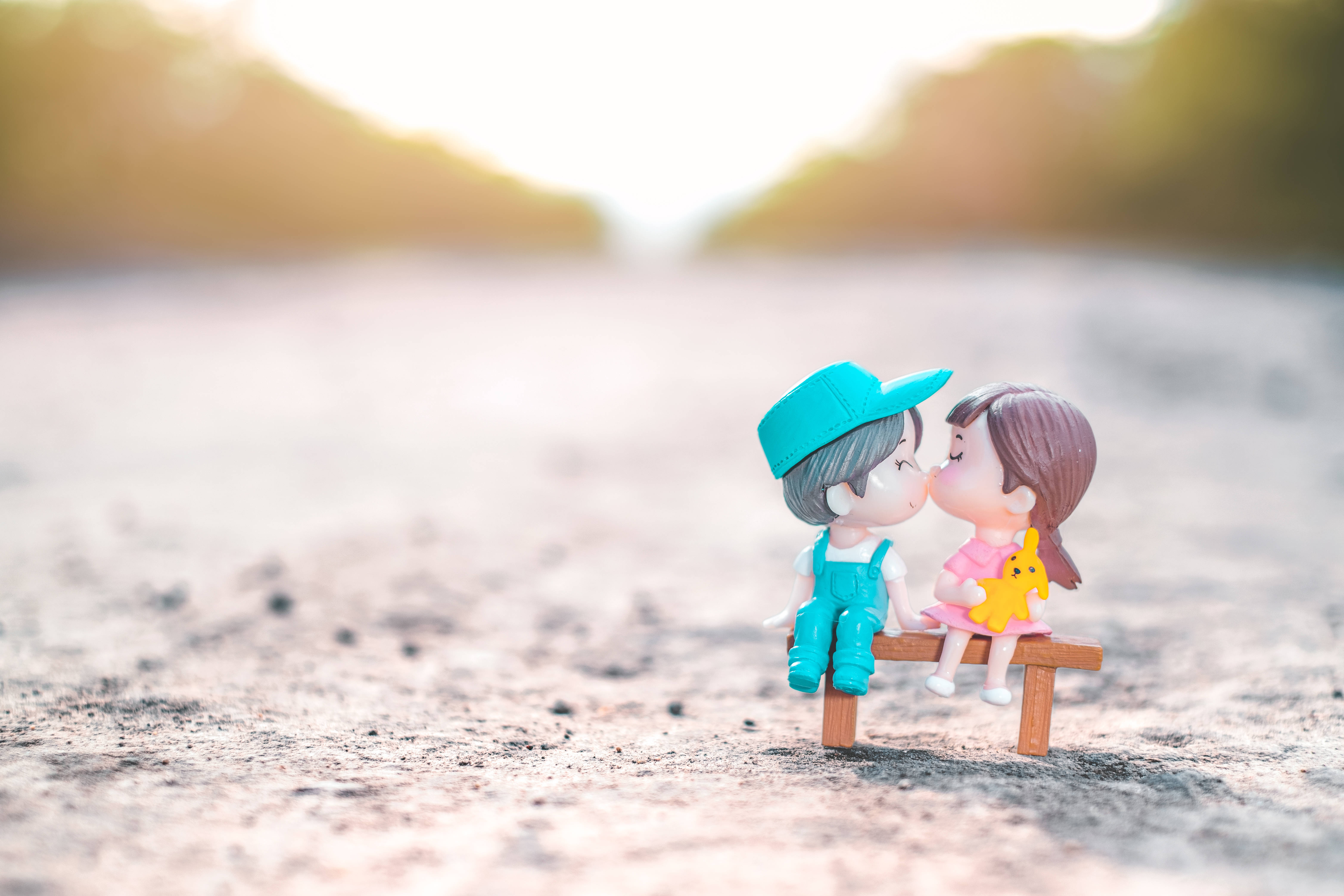 可愛粉紅女孩親吻 壁紙 — 兩個微型公仔喺溫暖嘅金色陽光下，喺一張細小嘅長凳上溫柔咁接吻。