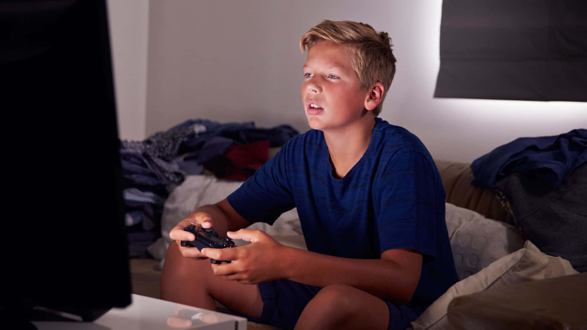 可愛美學青少年主機遊戲玩家 壁紙 — 一個專注的年輕男孩穿著藍色襯衫坐在沙發上，手持遊戲控制器，在昏暗的房間裡激烈地玩電子遊戲。