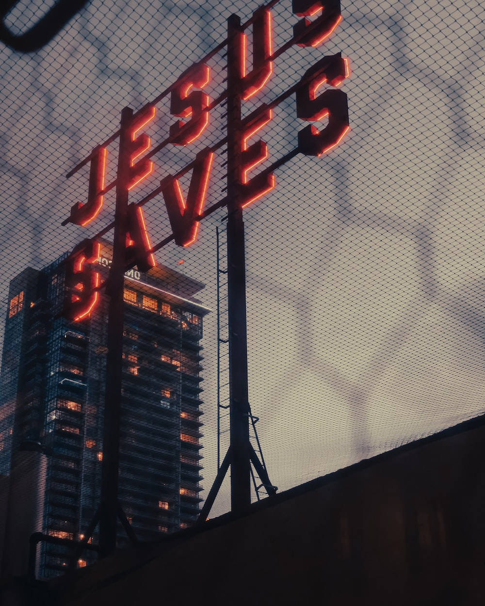 霓虹燈牌寫著 Jesus Saves，在圍欄後面，黃昏時分城市建築亮燈。