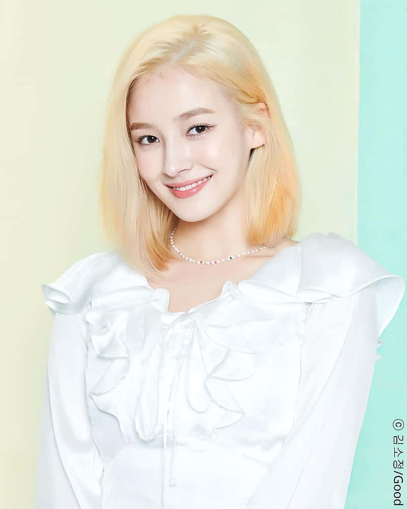 可愛韓國女團Momoland成員Nancy金髮白裙寫真 壁紙 — 一位微笑的年輕女性，留著金色波波頭，穿著白色荷葉邊襯衫，背景是柔和粉彩色。