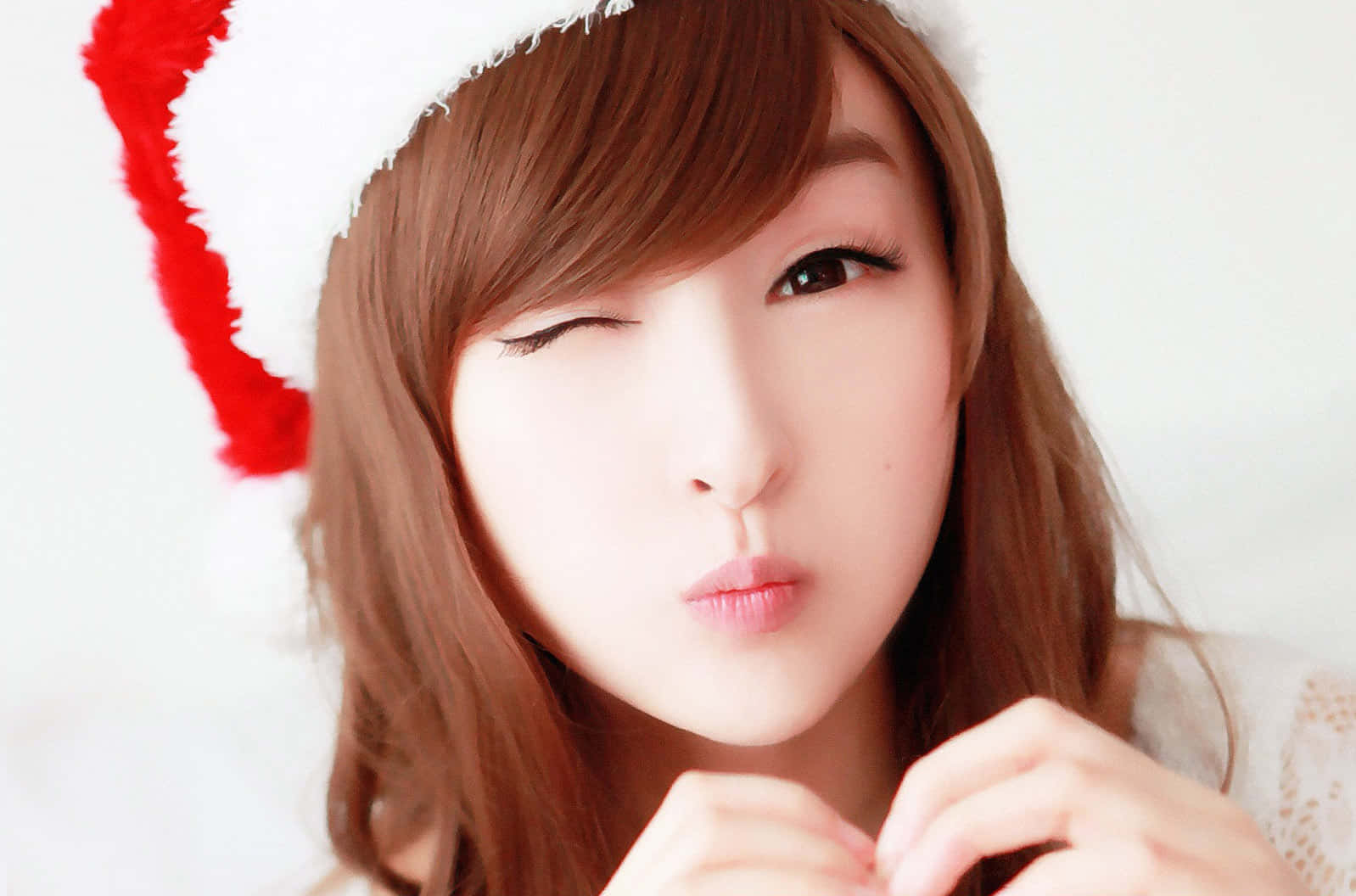 可愛韓國女孩戴聖誕帽 壁紙 — 一位棕色頭髮的年輕女性戴著紅白相間的聖誕帽，在明亮的白色背景前俏皮地眨眼。