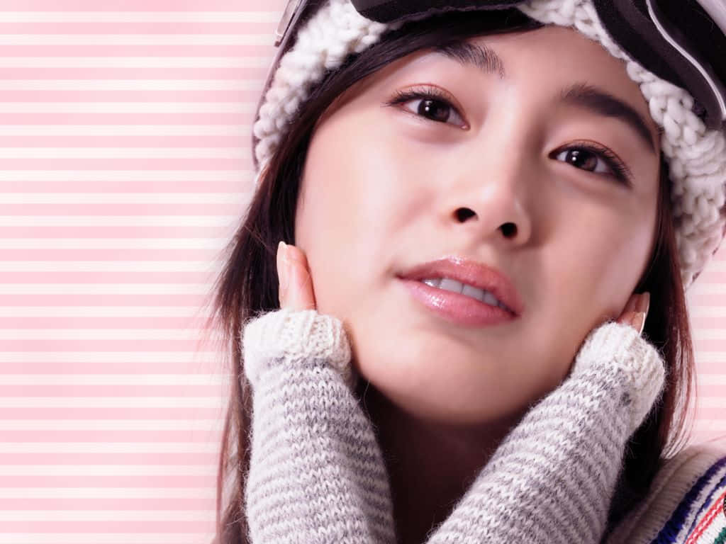 可愛韓國女演員金泰熙戴帽毛衣寫真 壁紙 — 一位穿著冬季裝備的年輕女性在粉紅色條紋背景上溫柔地微笑。
