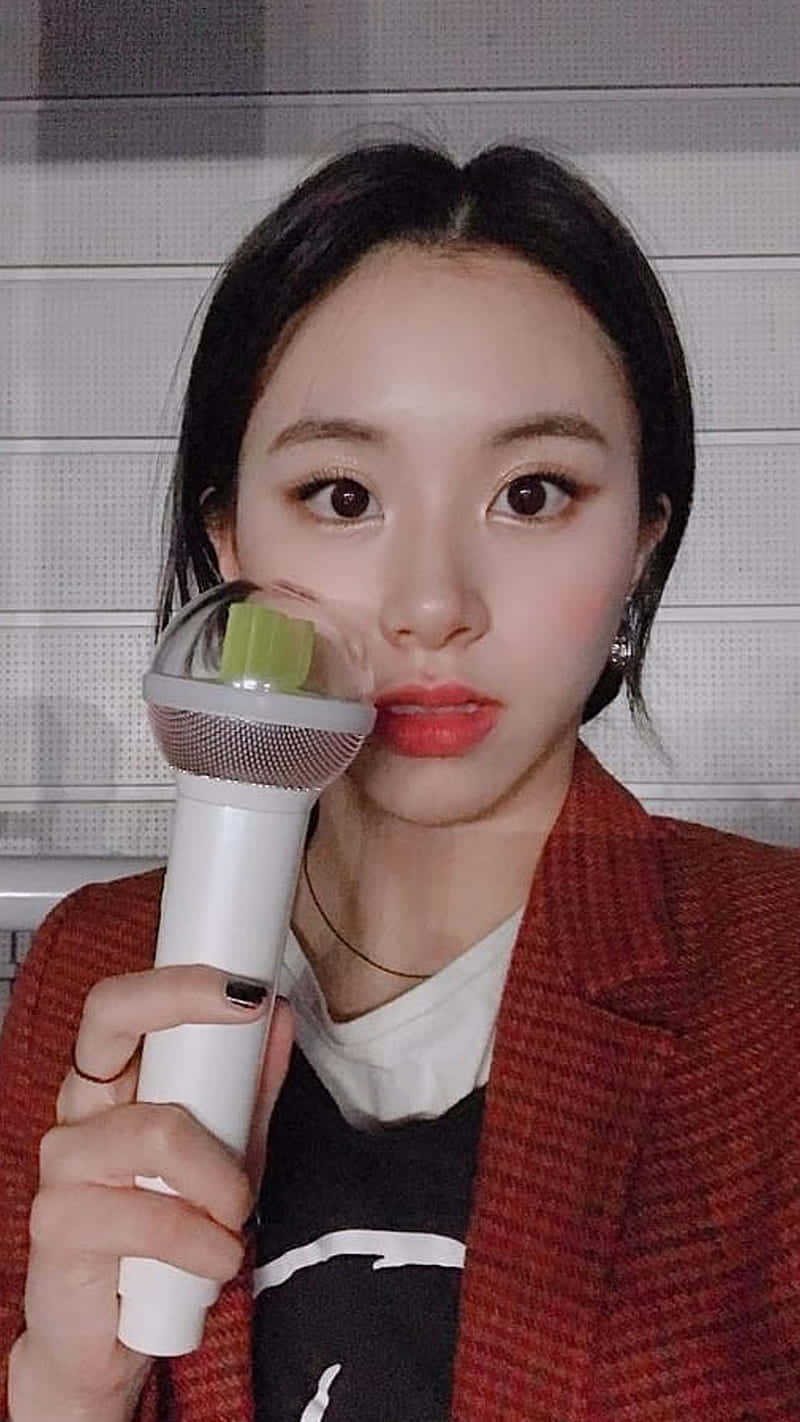 可愛韓國歌手Chaeyoung手持IU應援棒 壁紙 — 一位深色頭髮的年輕女性，在灰色瓷磚牆前將白色麥克風靠近臉部。