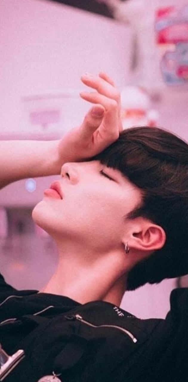 可愛韓國男孩黑衣靠粉牆暈倒姿勢寫真 壁紙 — 一位黑髮、身穿黑色外套的年輕男子，頭向後仰，閉著眼睛，手放在額頭上，背景是柔和的粉紅色。