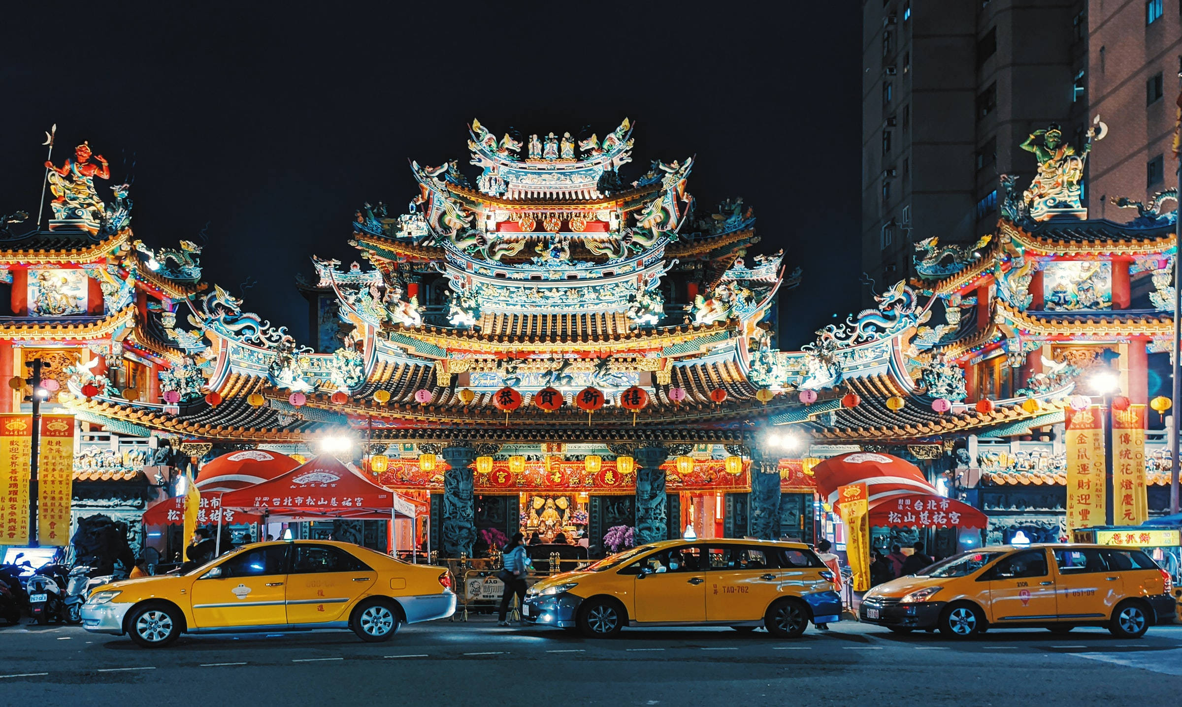 台北 Raohe 街夜景 壁紙 — 夜晚中璀璨燈光照耀的傳統寺廟，擁有華麗屋頂和紅色燈籠，兩旁是城市街道上的黃色計程車。