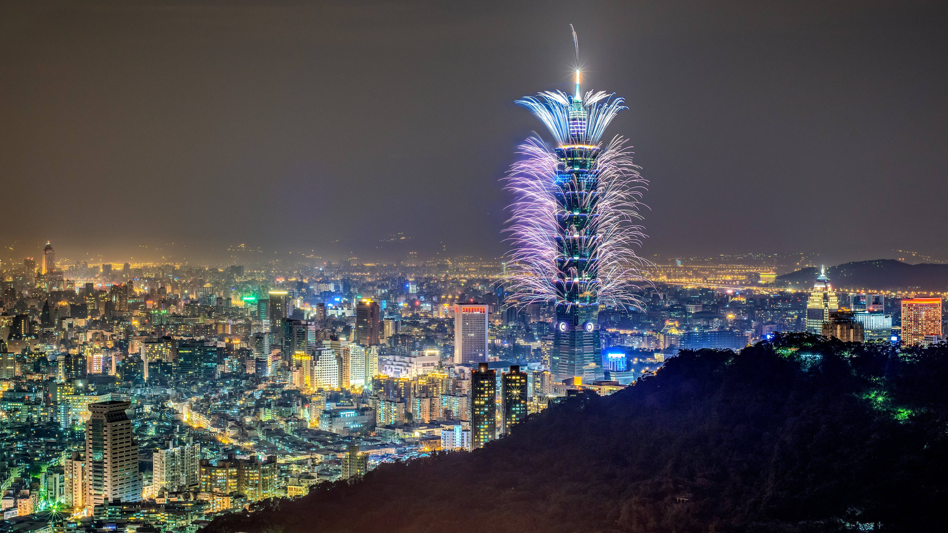 台北101台灣新年煙火 壁紙 — 煙火在台北101周圍爆炸，城市燈光照亮下方廣闊的城市天際線。