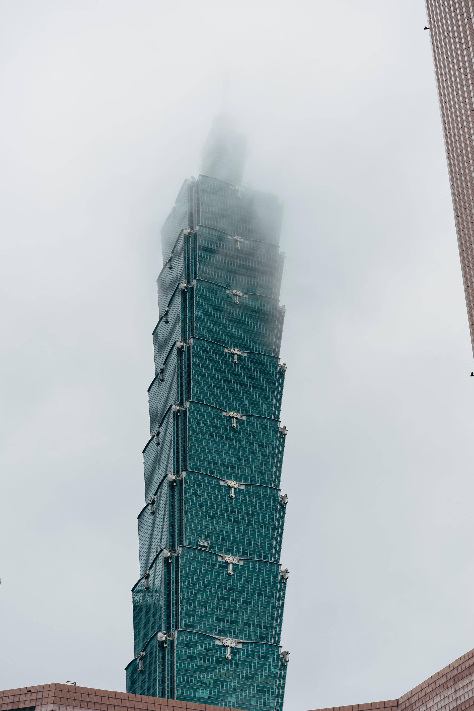 台北101霧氣景觀 壁紙 — 一座高聳的玻璃摩天大樓，部分被霧雲遮擋，映襯著灰色的天空。