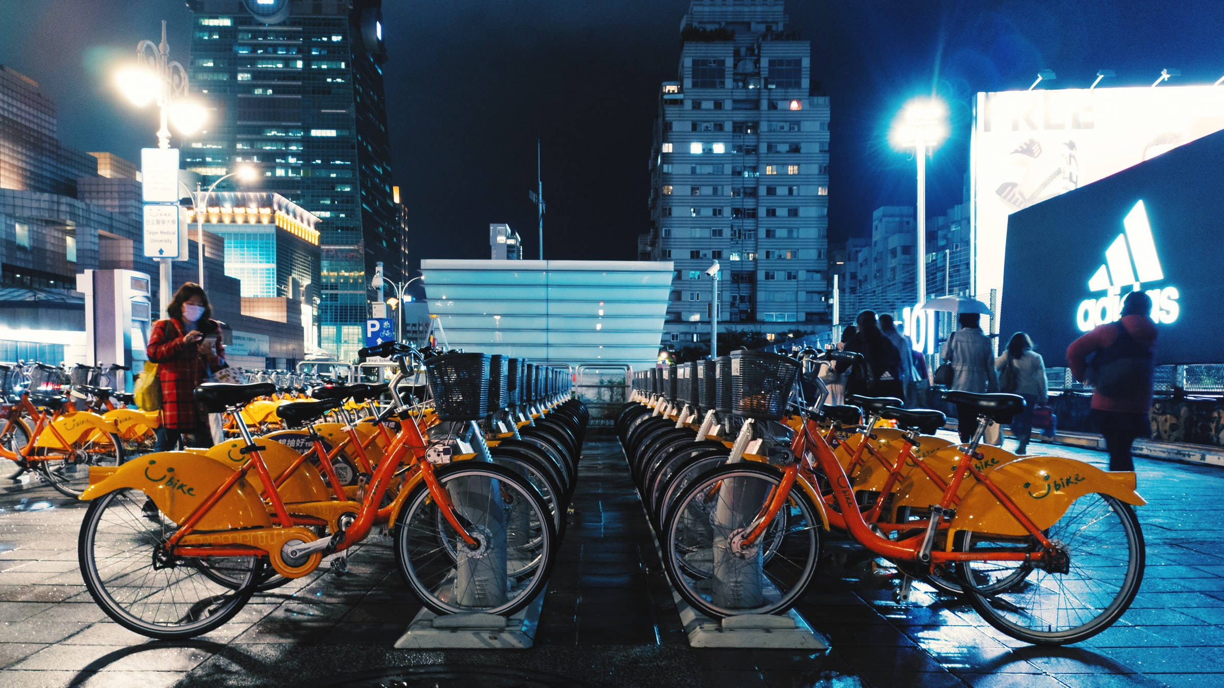 台北信義區自行車租賃站 壁紙 — 一排排亮橙色的共享單車在霓虹燈下閃爍，聳立的建築物和行人構成繁忙的夜景。