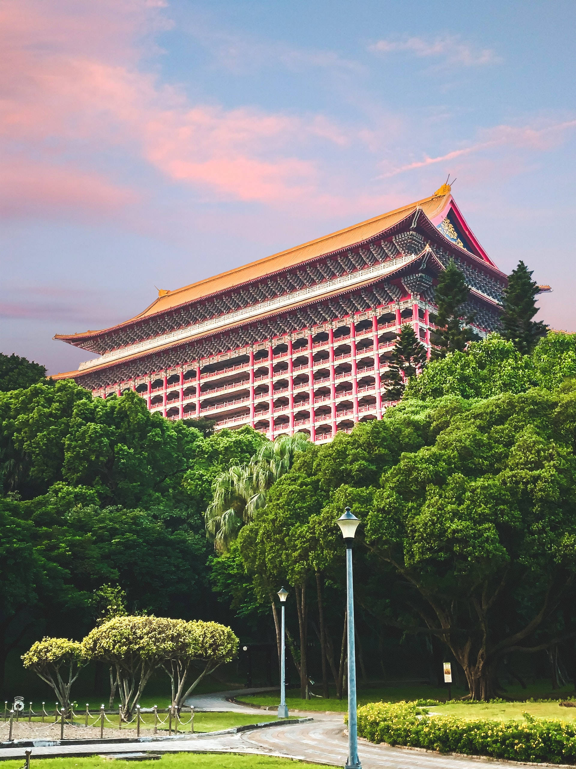 台北大飯店 壁紙 — 一座宏偉的傳統中式建築，擁有橙色屋頂和紅色陽台，在柔和的粉紅和藍色日落天空下，聳立於茂密的綠樹之上。