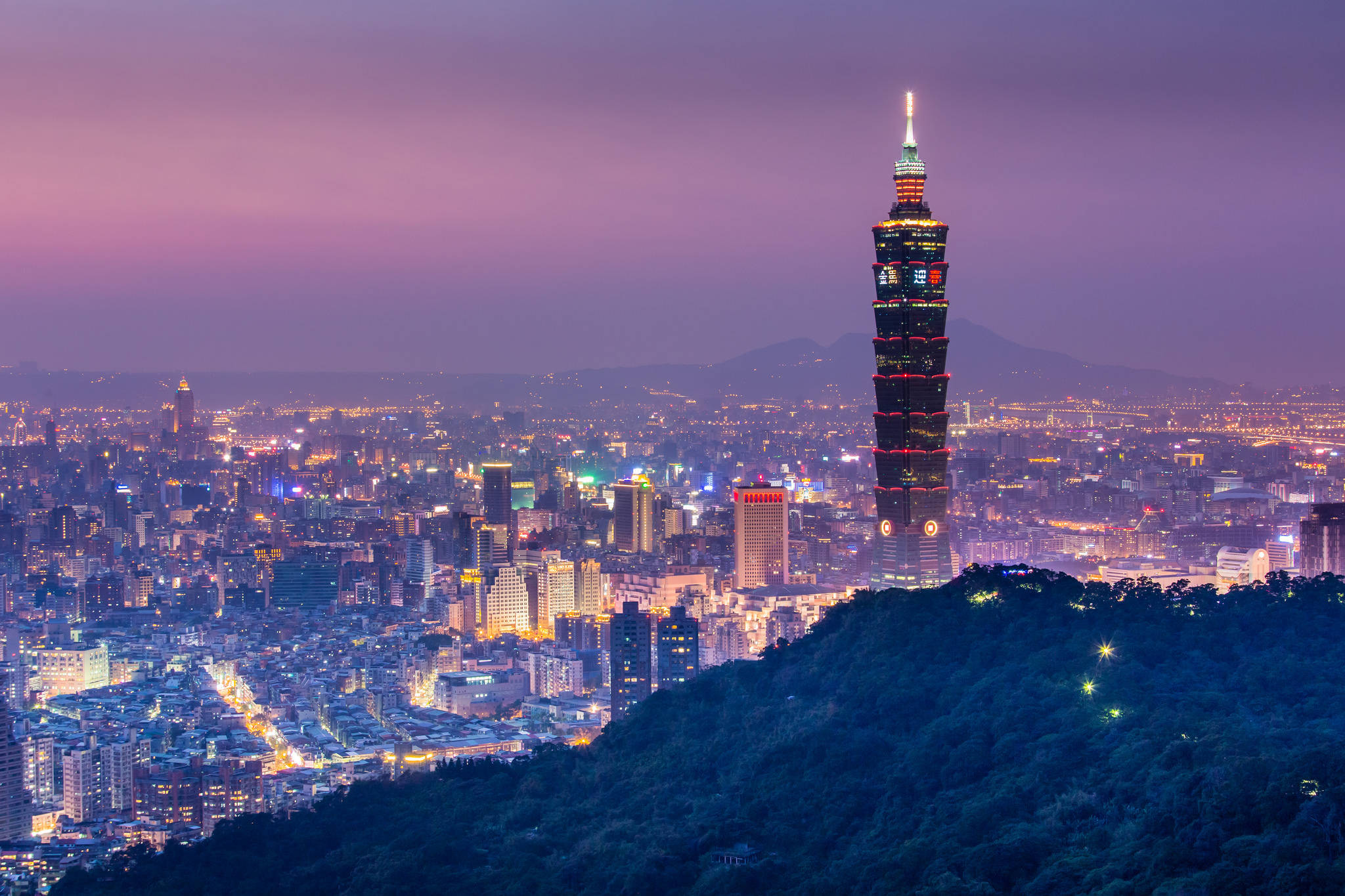 台北市台灣 壁紙 — 台北101摩天大樓在紫色暮色天空下，於群山環抱的廣闊城市景觀中閃耀。