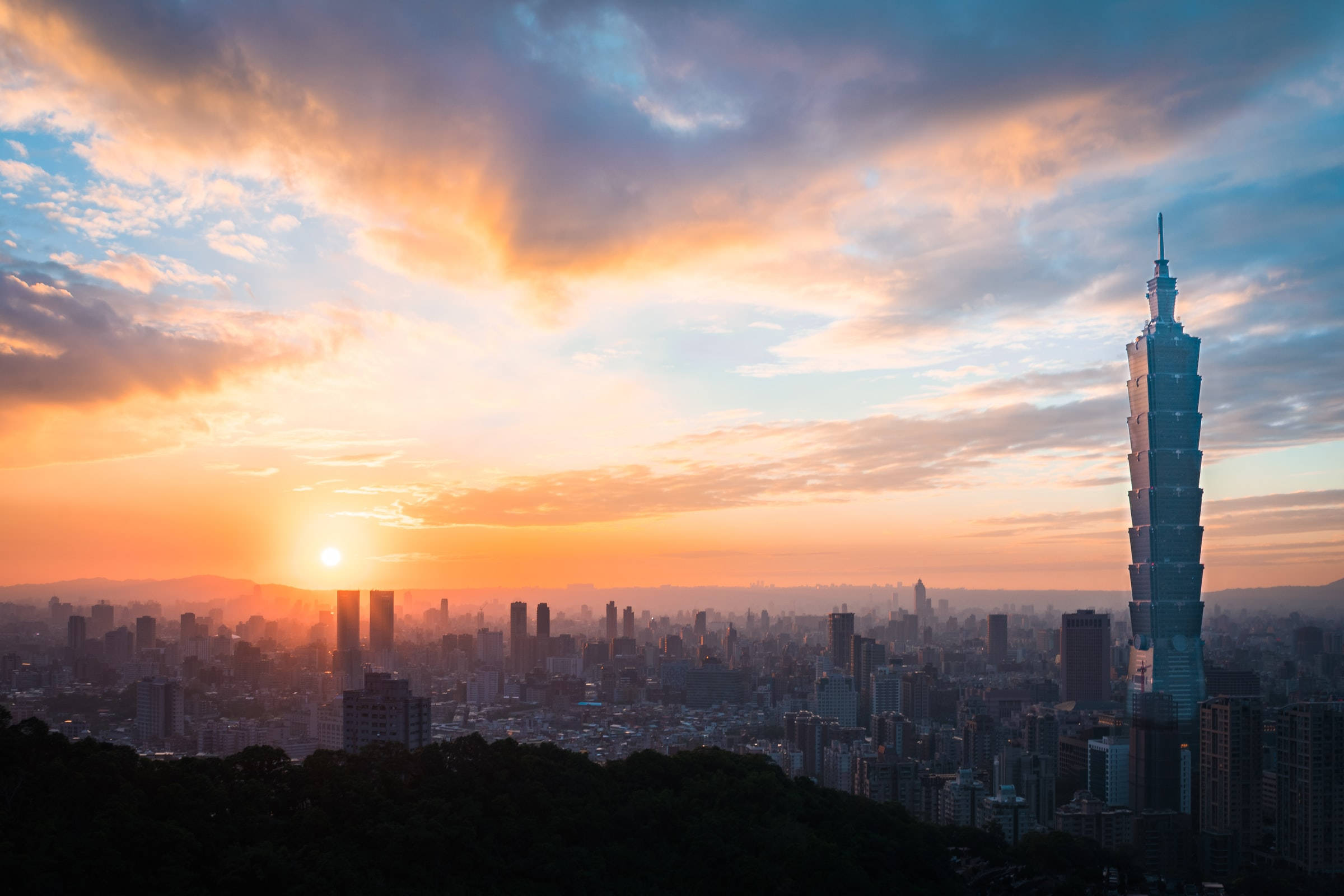 台北市夕陽景觀 壁紙 — 夕陽將台北天際線染成溫暖的橙色和藍色，台北101大樓在戲劇性的雲層中高聳入雲。