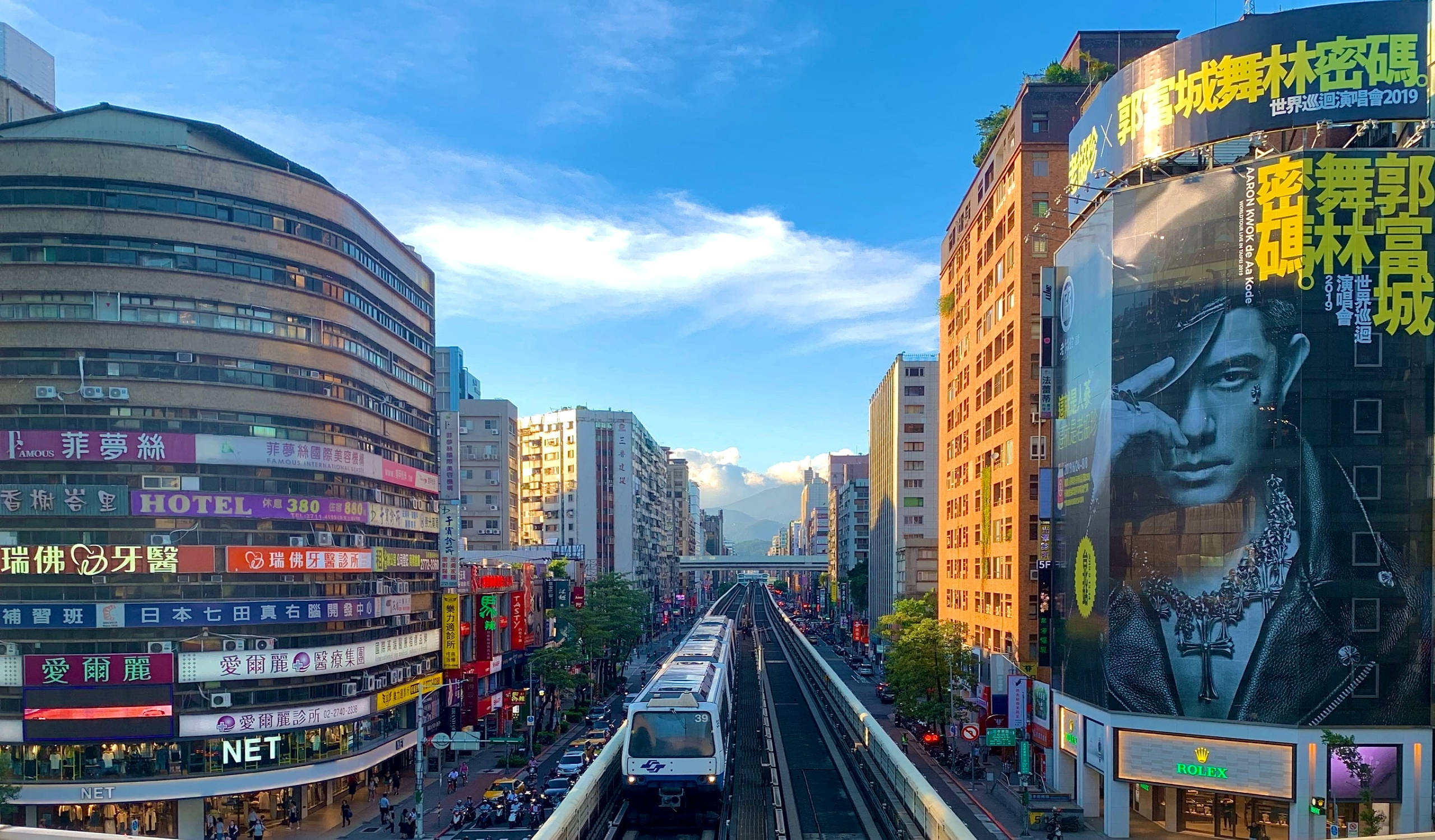 台北市高架捷運與天際線 壁紙 — 充滿活力的城市街景，單軌列車在高樓大廈之間穿梭，天空湛藍，白雲點綴。