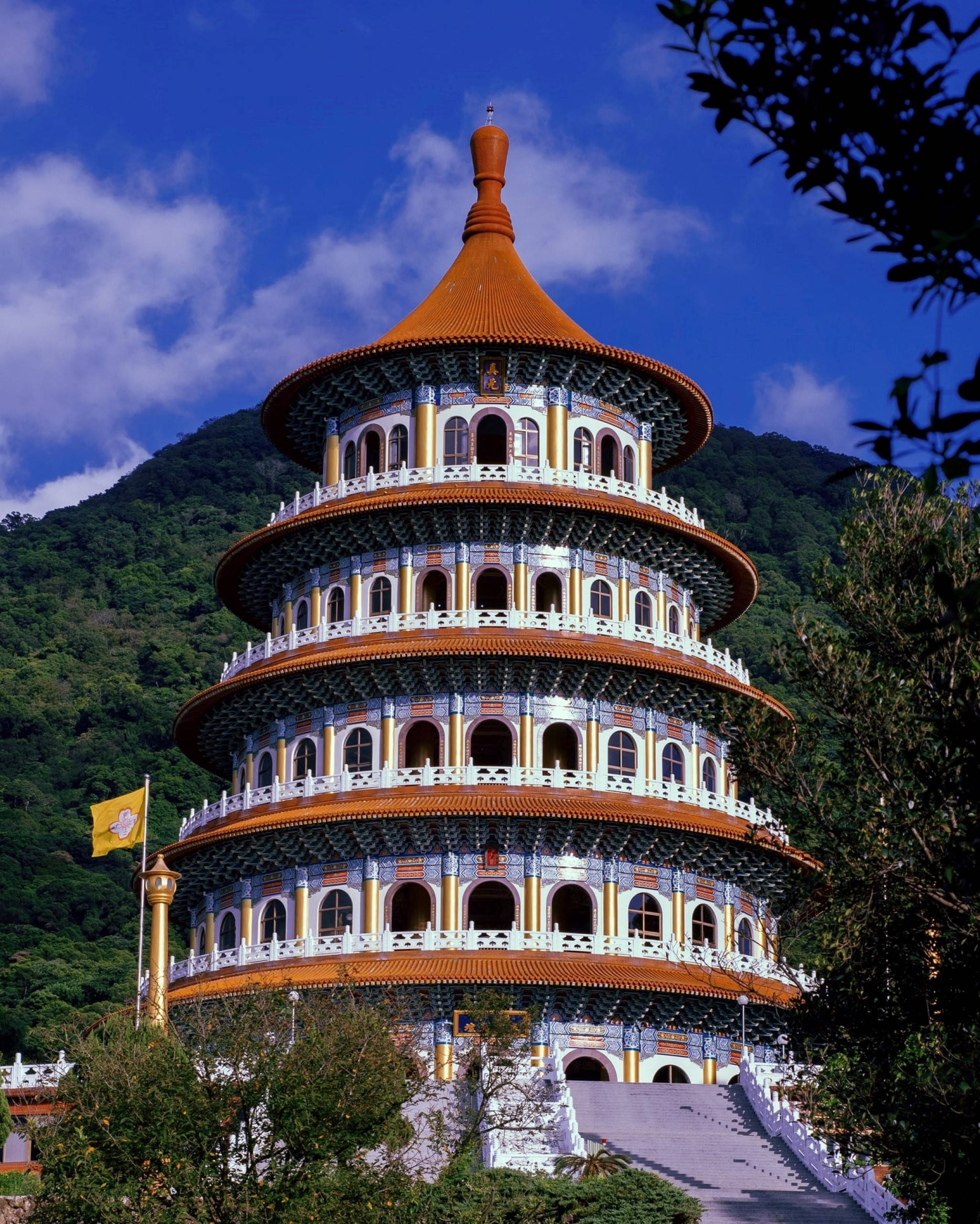 台北新市區無極天元宮 壁紙 — 一座宏偉的多層寺廟，擁有橙色屋頂和白色欄杆，在明亮的藍天下雄偉地矗立在鬱鬱蔥蔥的綠色山巒前。