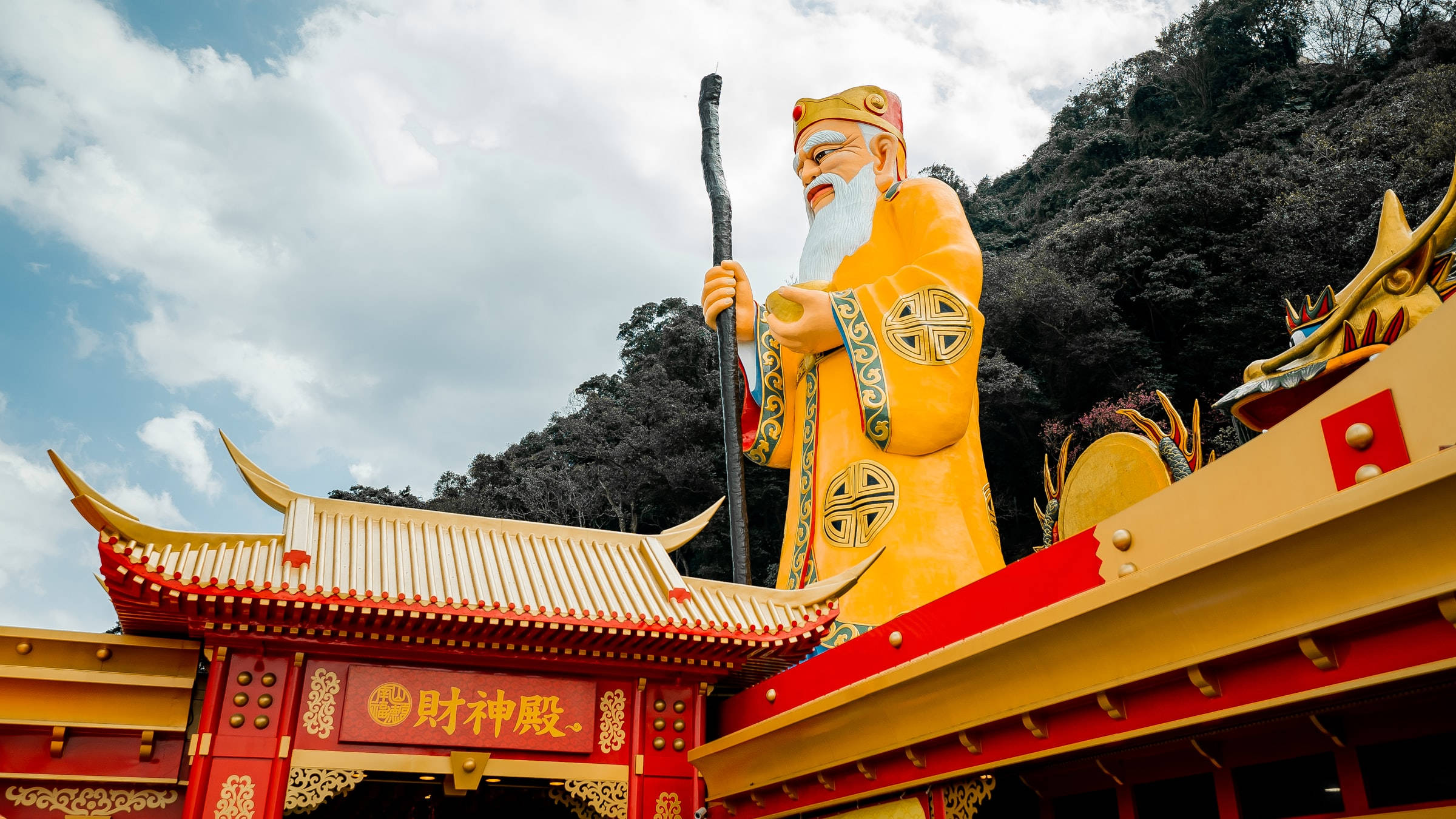 台北紅樓南山福德祠 壁紙 — 一座高聳的金色鬍鬚神祇雕像手持權杖，矗立在色彩鮮豔的紅金色寺廟頂上，背景是多雲的天空。
