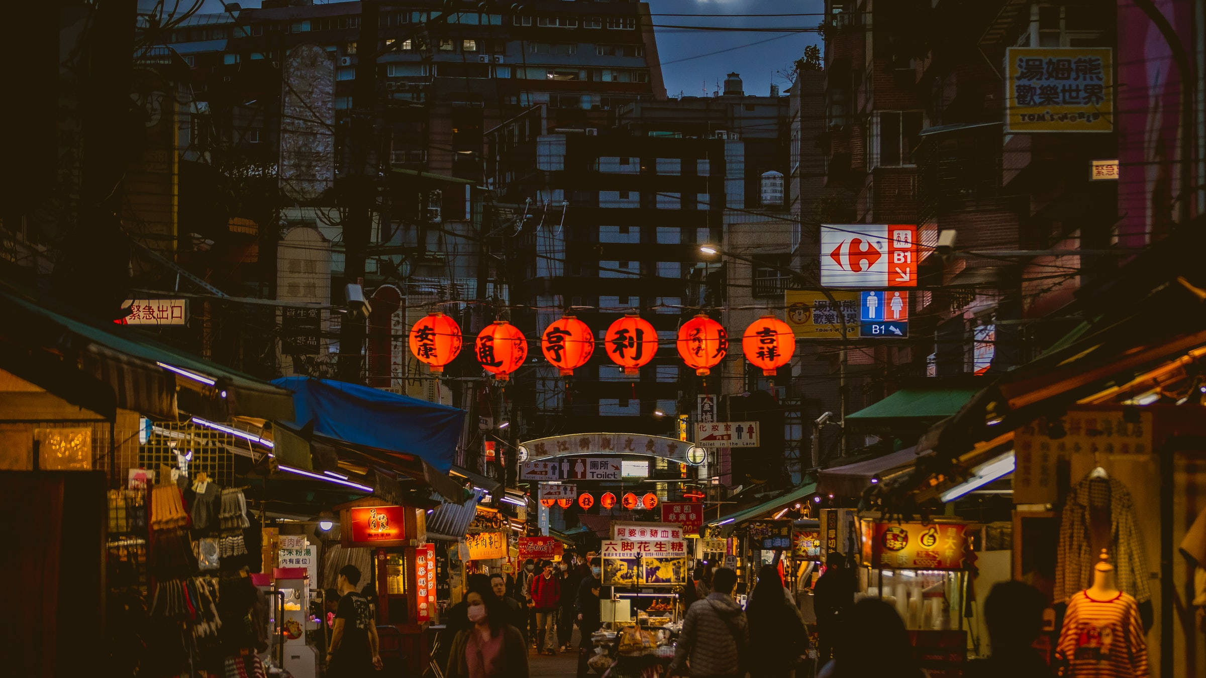 台北街燈夜景 壁紙 — 一條熱鬧的夜市小巷，被發光的紅燈籠和霓虹燈招牌照亮，襯托著高聳的城市建築。