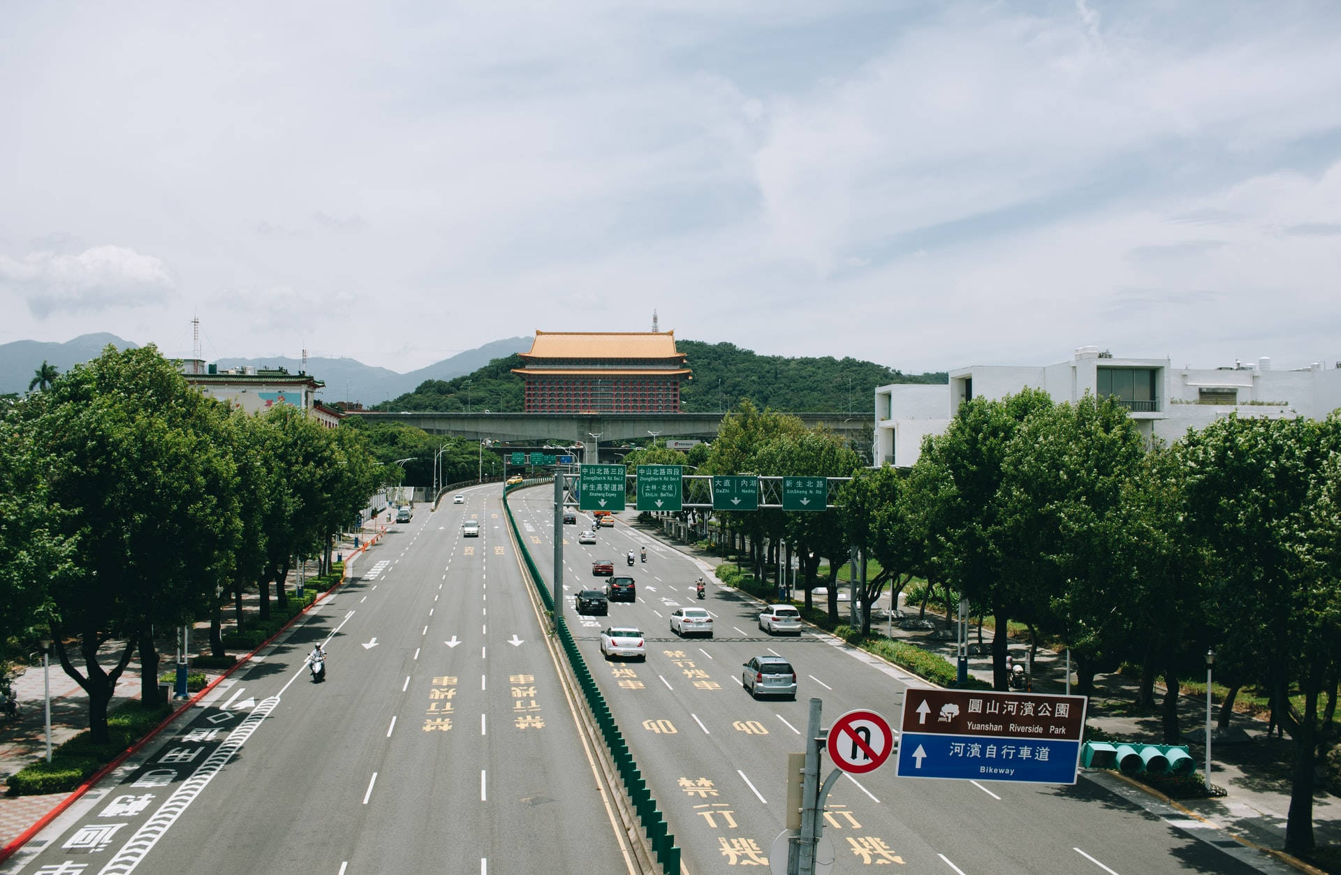 台北通往世貿公園的高速公路 壁紙 — 寬闊的城市高速公路兩旁綠樹成蔭，通向山頂上一座傳統中式建築，天空多雲。