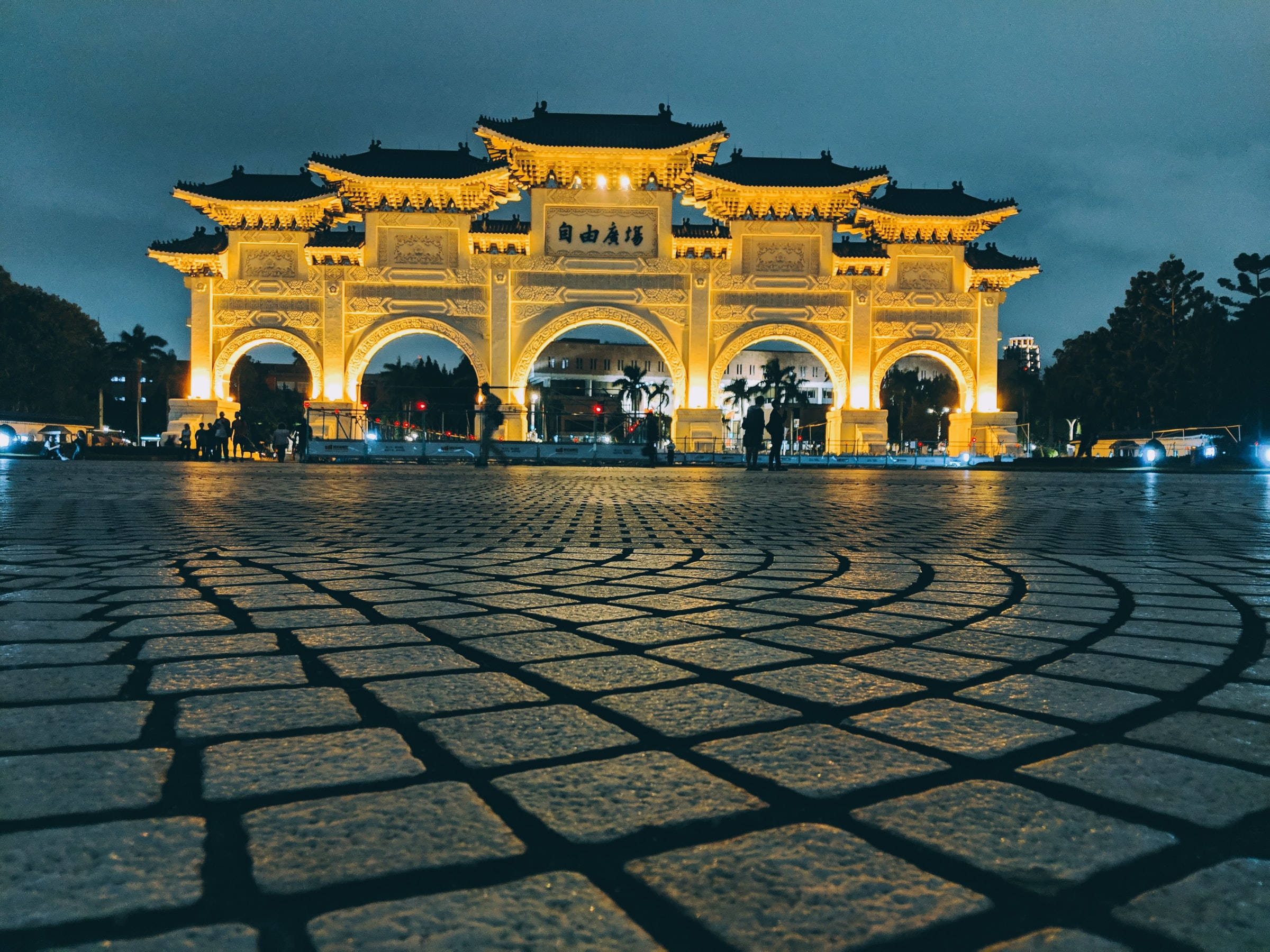 台北金色拱門 壁紙 — 一座宏偉的照明傳統中式牌坊在黃昏時分溫暖地發光，在濕漉漉的鵝卵石廣場上投下長長的影子。