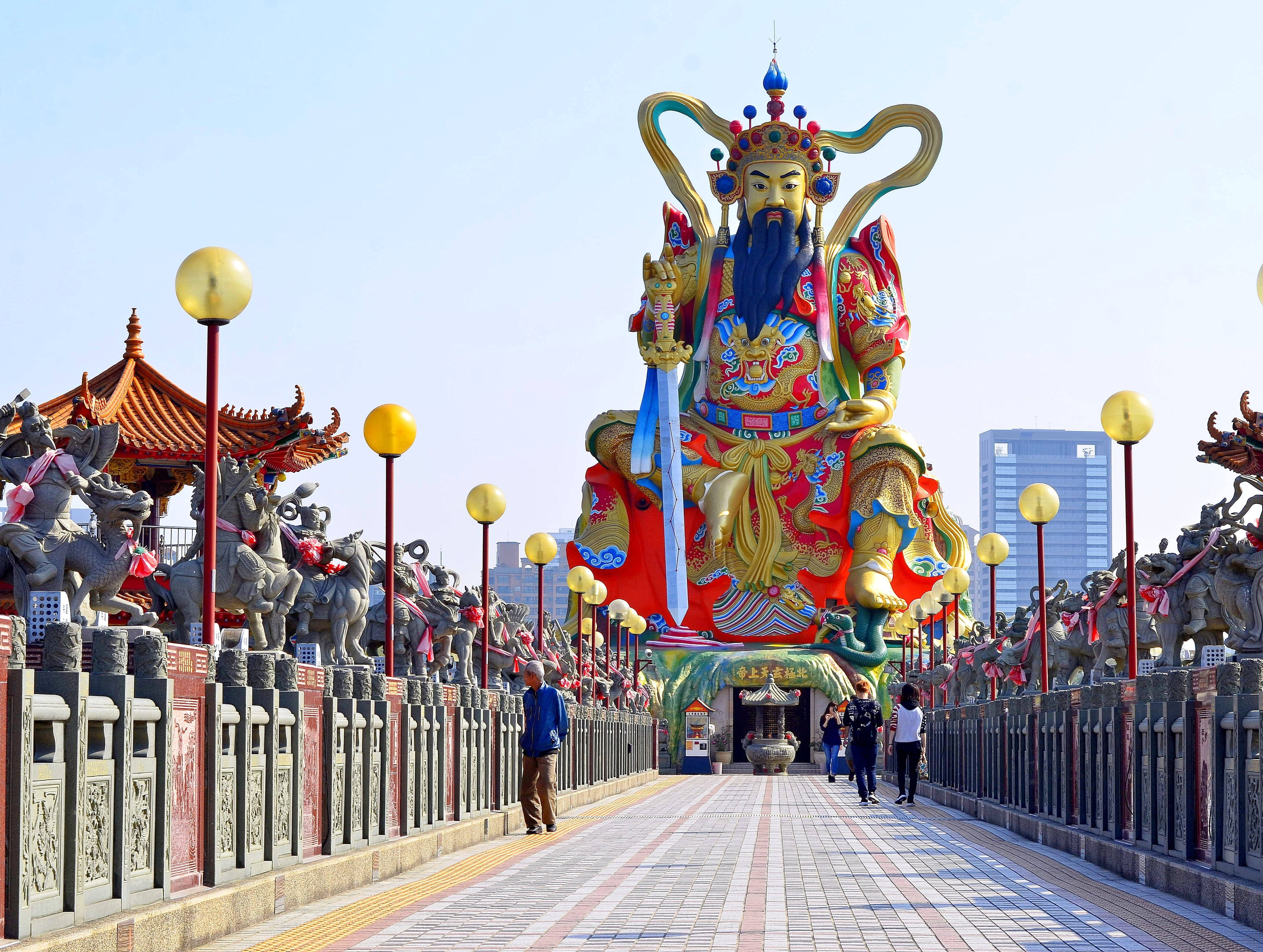 台灣上帝雕像 壁紙 — 一座鬍鬚神祇的宏偉色彩鮮豔雕像，坐落在石獅守護和黃色燈籠點綴的橋上，背景是清澈的藍天。