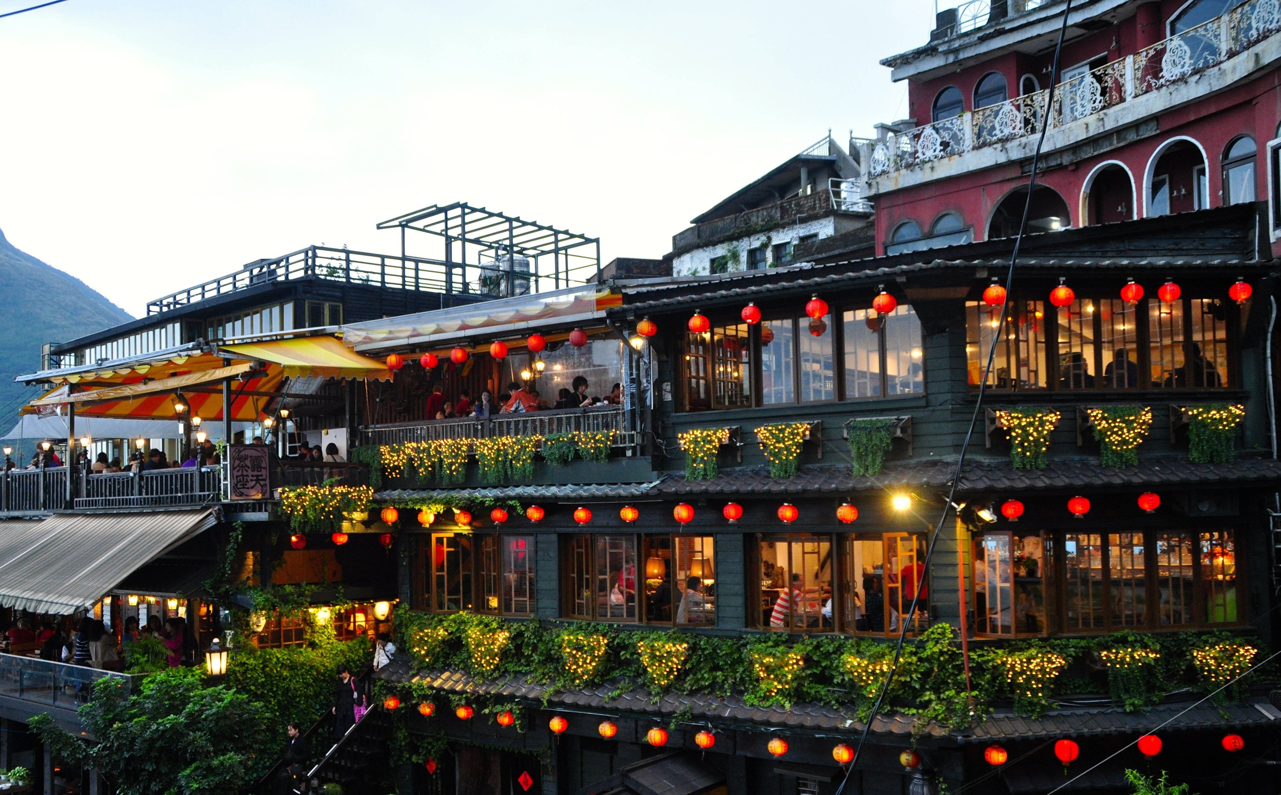 台灣九份阿妹茶館 壁紙 — 黃昏時分，一個充滿活力的山坡小鎮，紅色燈籠閃爍，綠意盎然，點綴著多層建築，裡面擠滿了人。