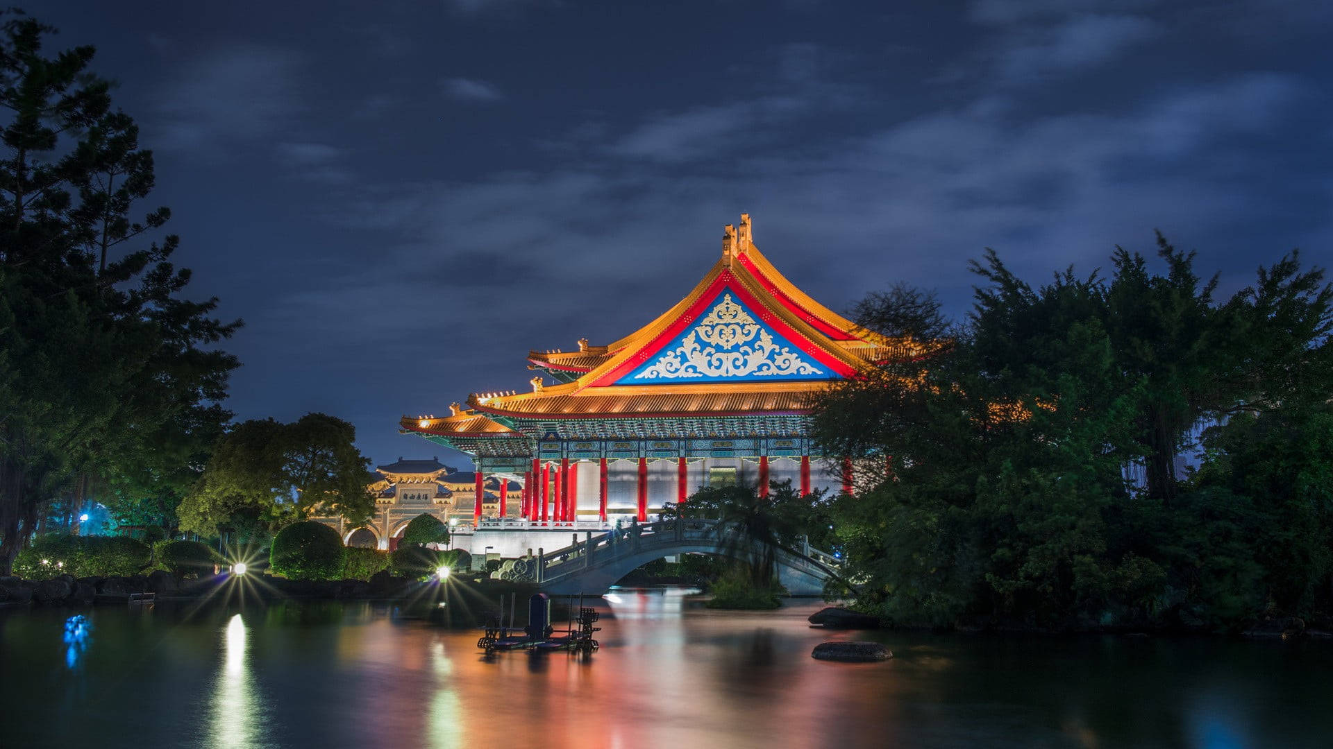 台灣國家戲劇院 壁紙 — 一座宏偉的傳統中式亭閣，帶有發光的紅色和金色屋頂燈光，在漆黑的夜空下美麗地倒映在平靜的水面上。