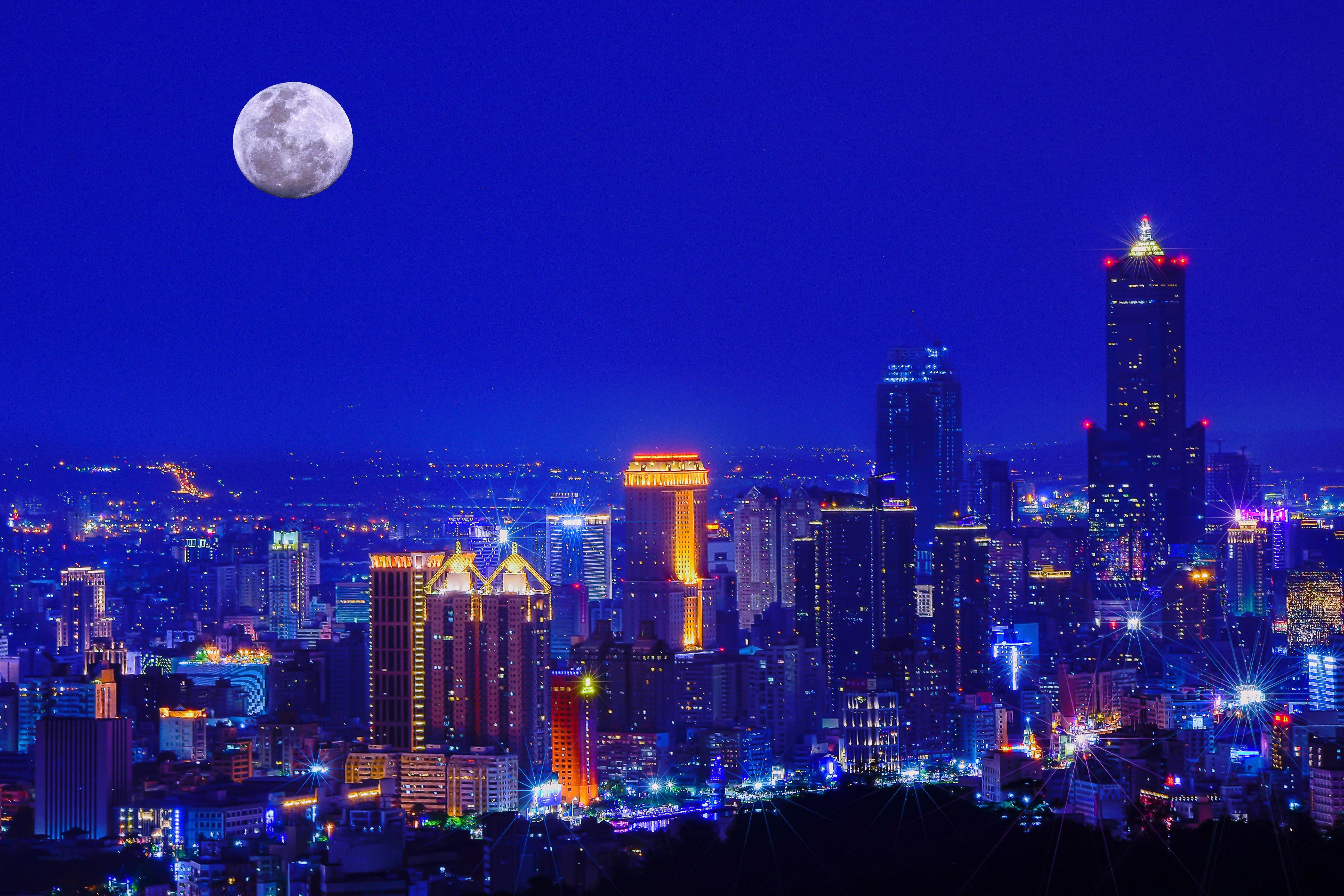 台灣城市夜景 壁紙 — 在深藍色天空下，一個充滿活力的城市天際線在夜晚被五彩繽紛的燈光照亮，一輪巨大的滿月高懸其上。