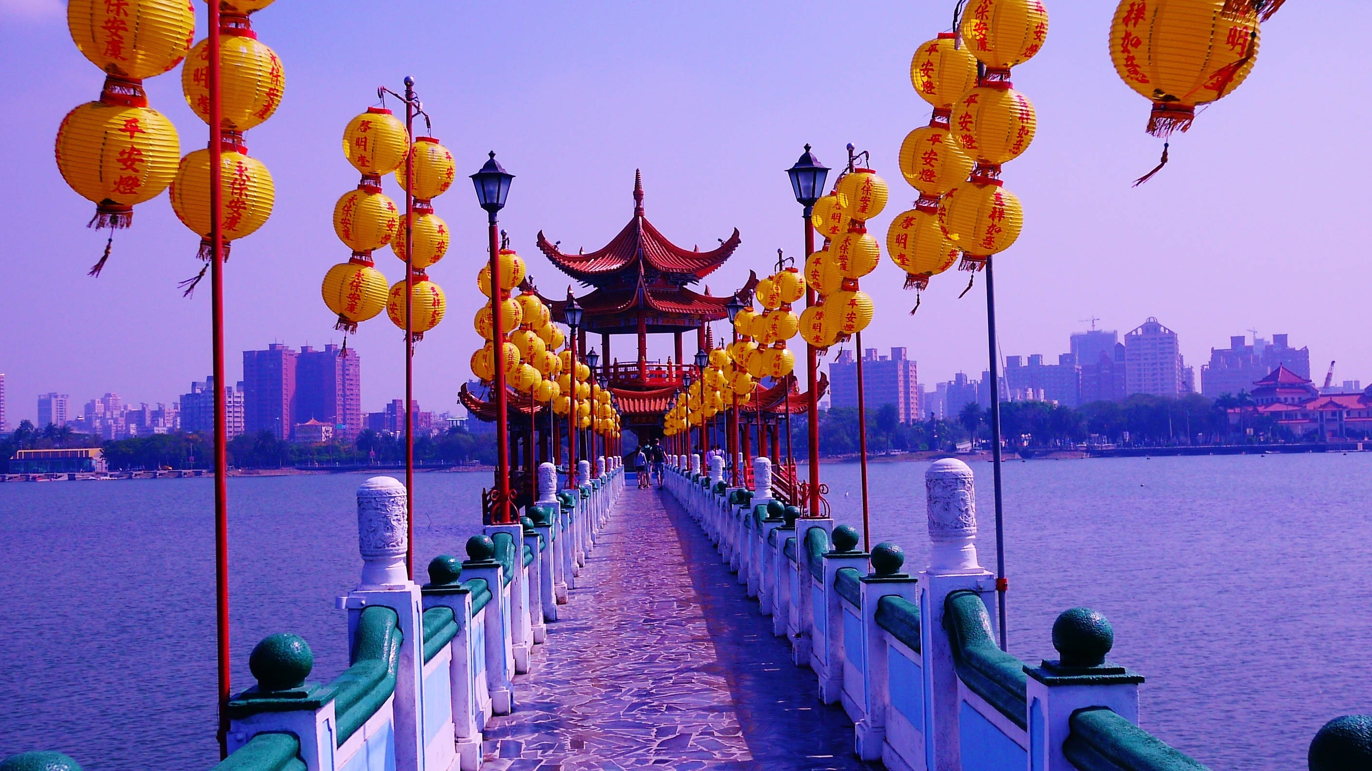 台灣春秋閣 壁紙 — 一座充滿活力的橋樑，裝飾著發光的黃色燈籠，橫跨平靜的水面，通往一座傳統亭閣，背景是朦朧的城市天際線。