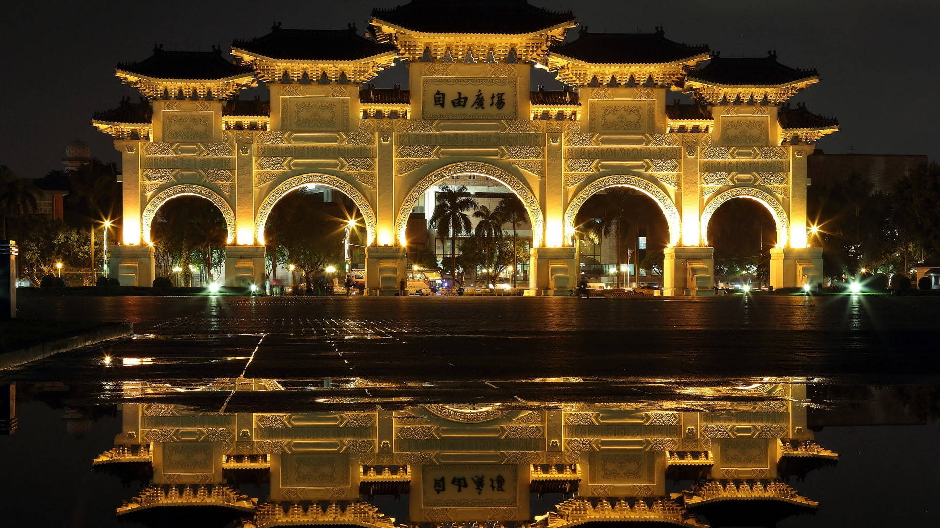 台灣自由廣場拱門 壁紙 — 一座宏偉的傳統中式牌坊在夜晚被照亮，其金色燈光完美地倒映在下方濕漉漉的廣場上。
