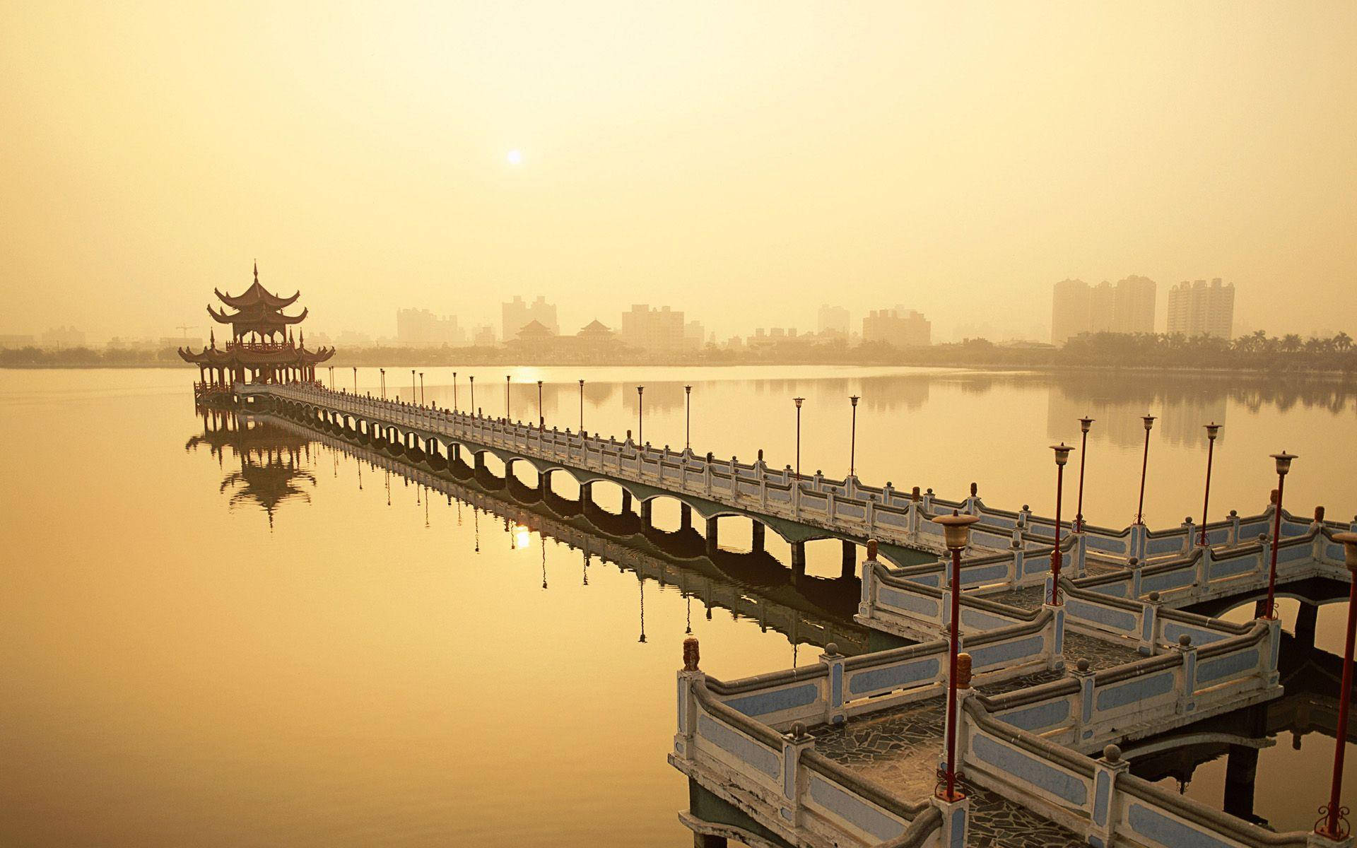 台灣蓮花池湖景 壁紙 — 一條長長的華麗橋樑，上面有傳統亭閣，在日出時延伸過平靜的金色水面，朝向朦朧的城市天際線。