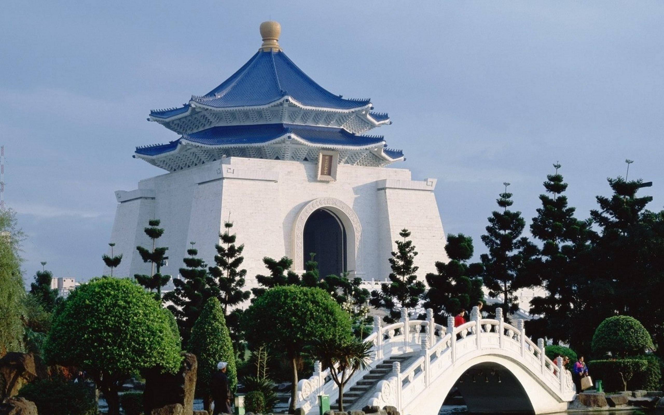 台灣蔣中正紀念堂壯觀影像 壁紙 — 一座巨大的白色紀念碑，藍色瓦片屋頂，矗立在一座白色拱橋後面，周圍是修剪整齊的綠植，天空晴朗。
