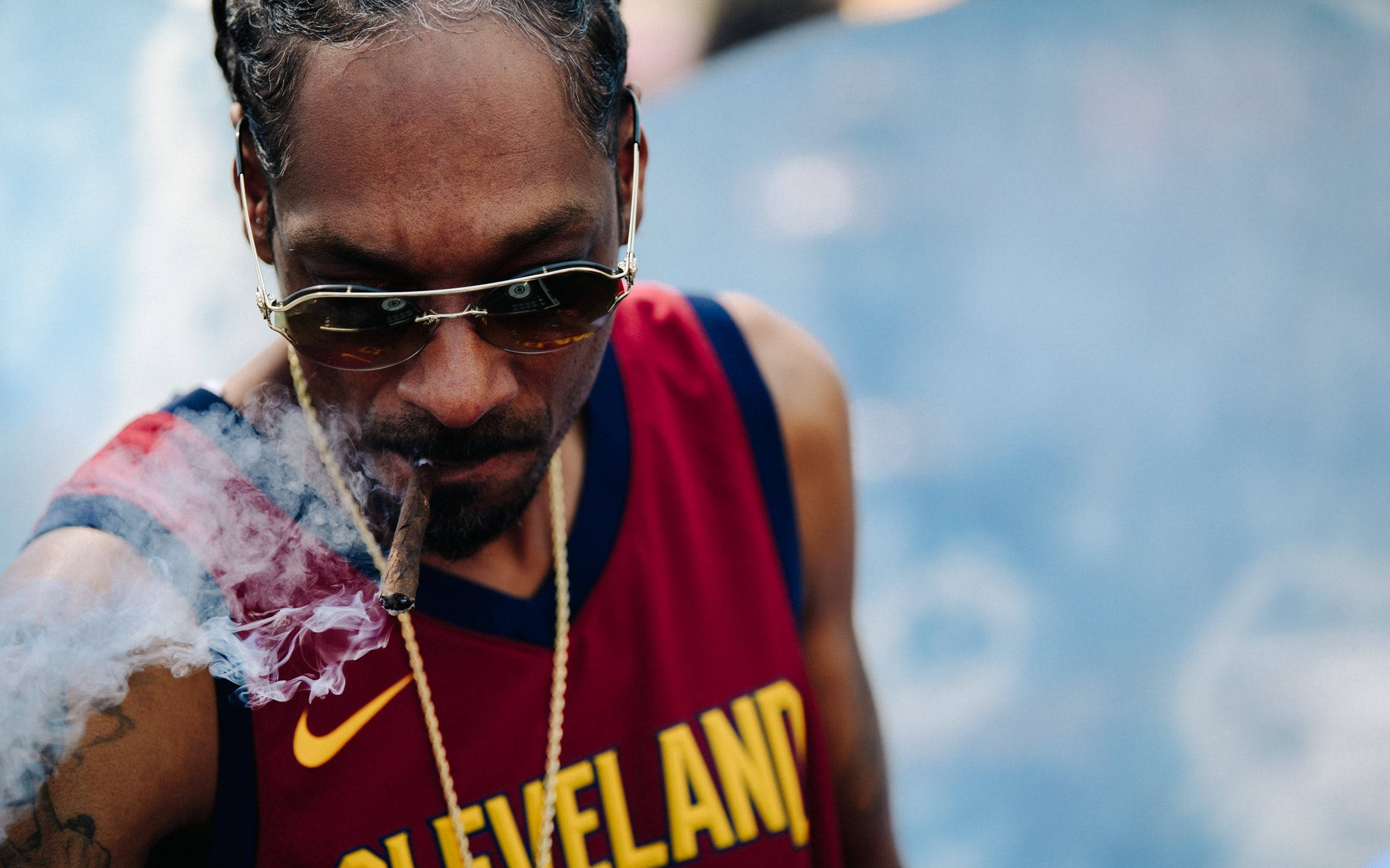 史努比狗狗穿克里夫蘭球衣 壁紙 — Snoop Dogg 喺模糊嘅藍色背景下，着住紅色克里夫蘭波衫食雪茄，煙霧喺佢塊面周圍打轉。