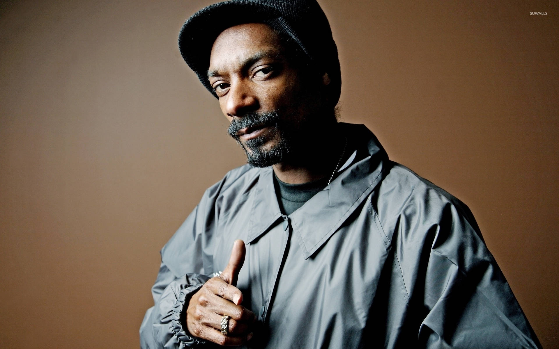 史努比狗狗身穿灰色襯衫 壁紙 — Snoop Dogg 穿著灰色外套和黑色冷帽，在溫暖的棕色背景下自信地豎起大拇指。