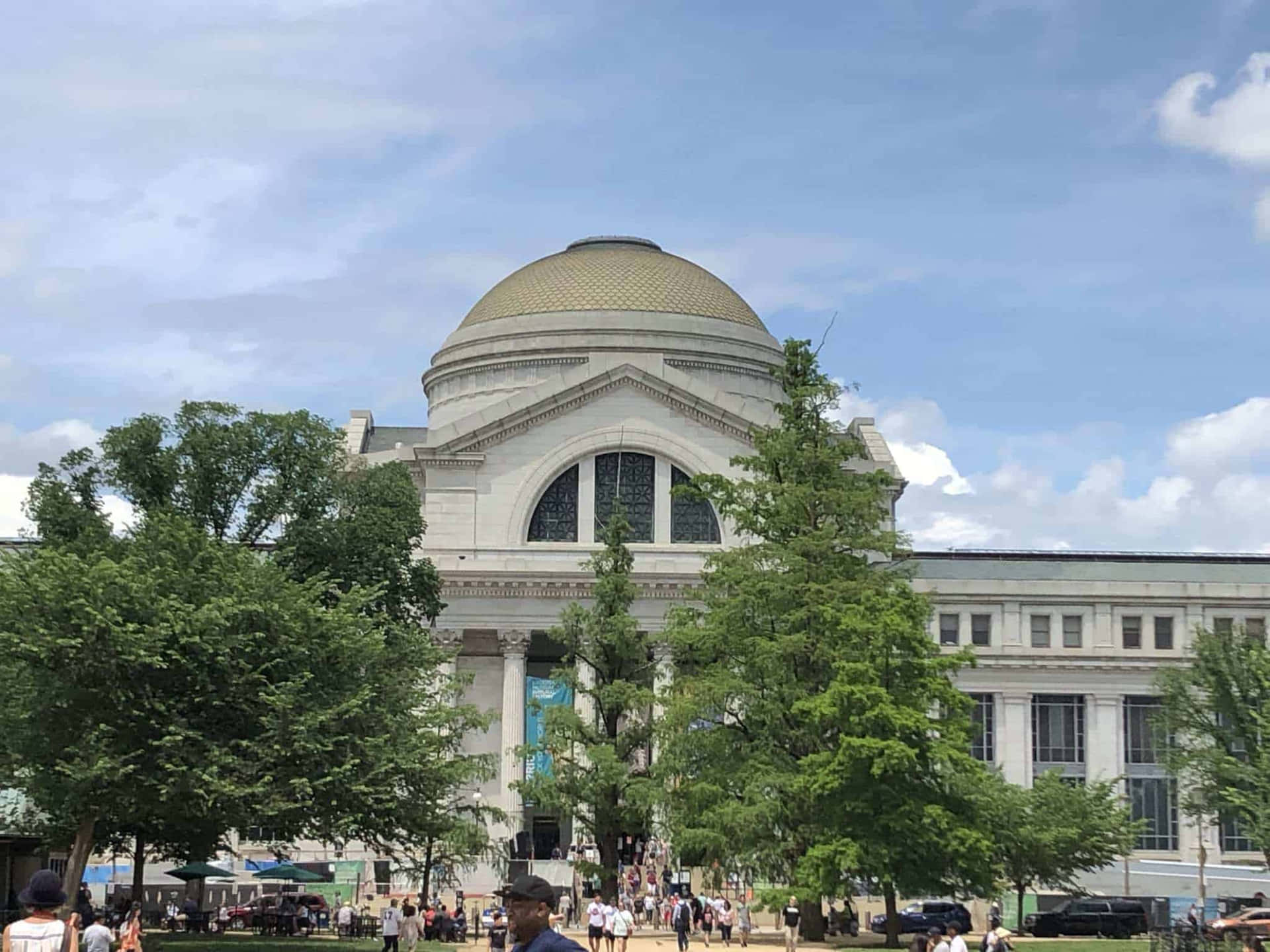 宏偉的白色建築，金色圓頂，周圍有綠樹和人們在陽光明媚的日子裡。